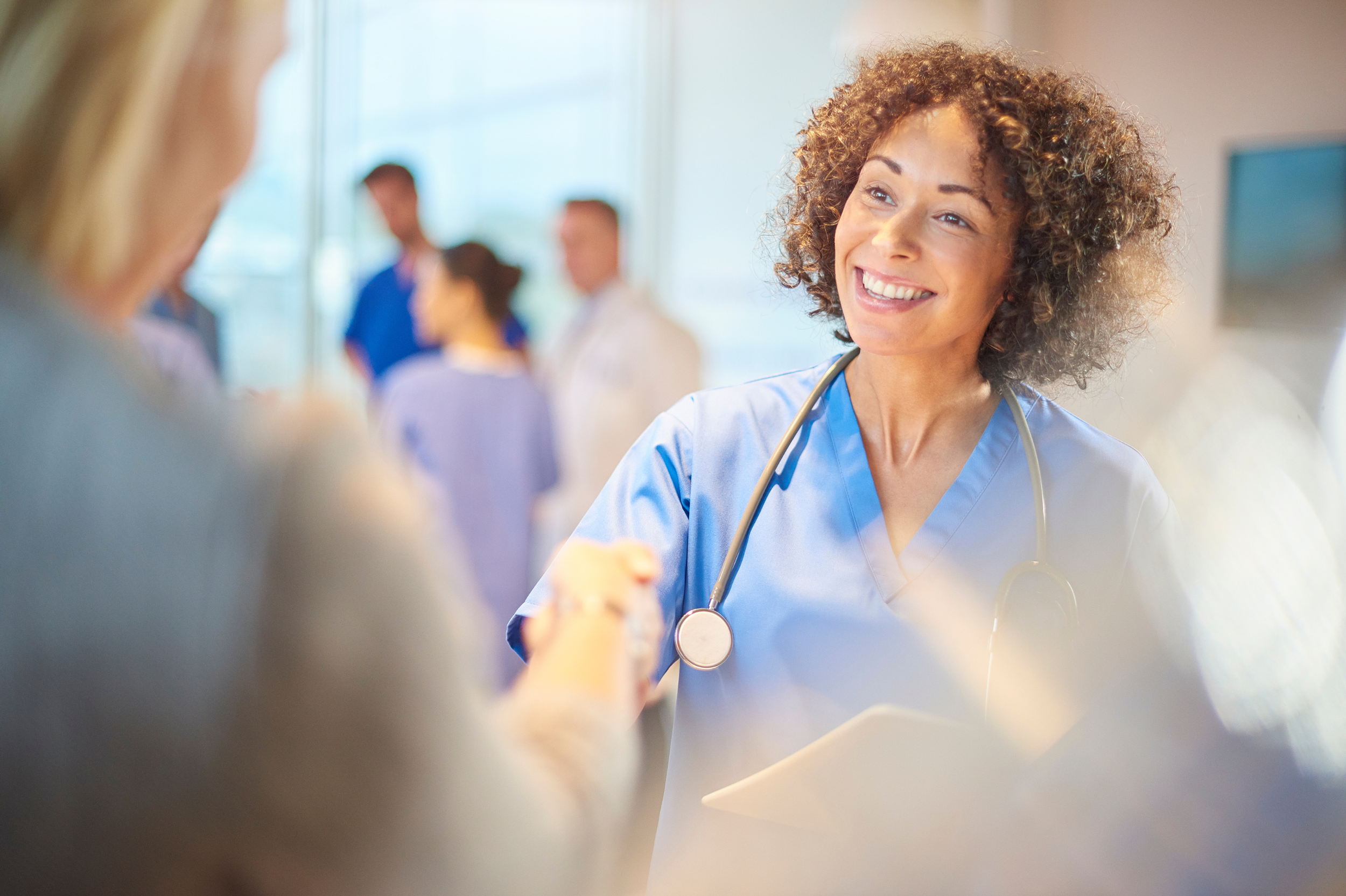 A smiling female healthcare professional in blue scrubs with a stethoscope around her neck, shaking hands with another person in a busy hospital or medical setting.