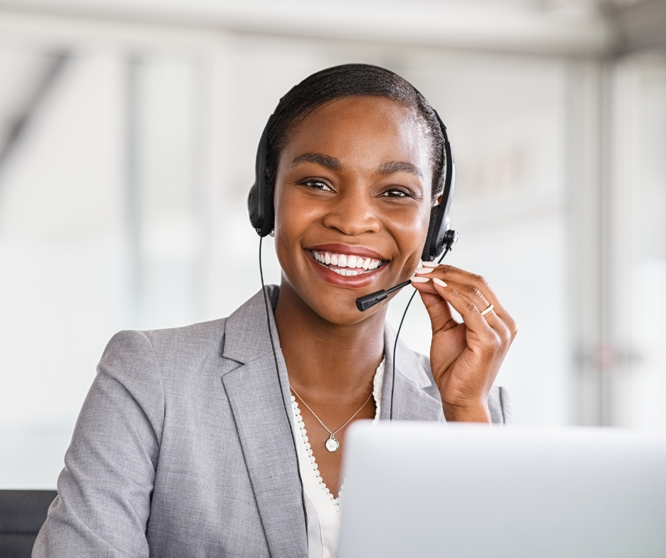 A smiling woman wearing a headset, sitting at a desk with a laptop in a modern office setting.