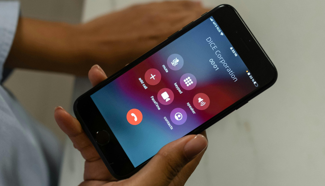 A person holding a smartphone in their left hand and on a phone call with DICE Corporation. The call duration is 1 second. The phone interface shows options for mute, keypad, speaker, add call, faceTime, contacts, and end call. The background wall is light-colored.
