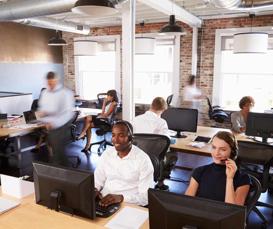 Office environment with multiple employees working at their desks. Some are wearing headsets, indicating customer service or call center roles. The office has large windows, exposed brick walls, and modern lighting fixtures.