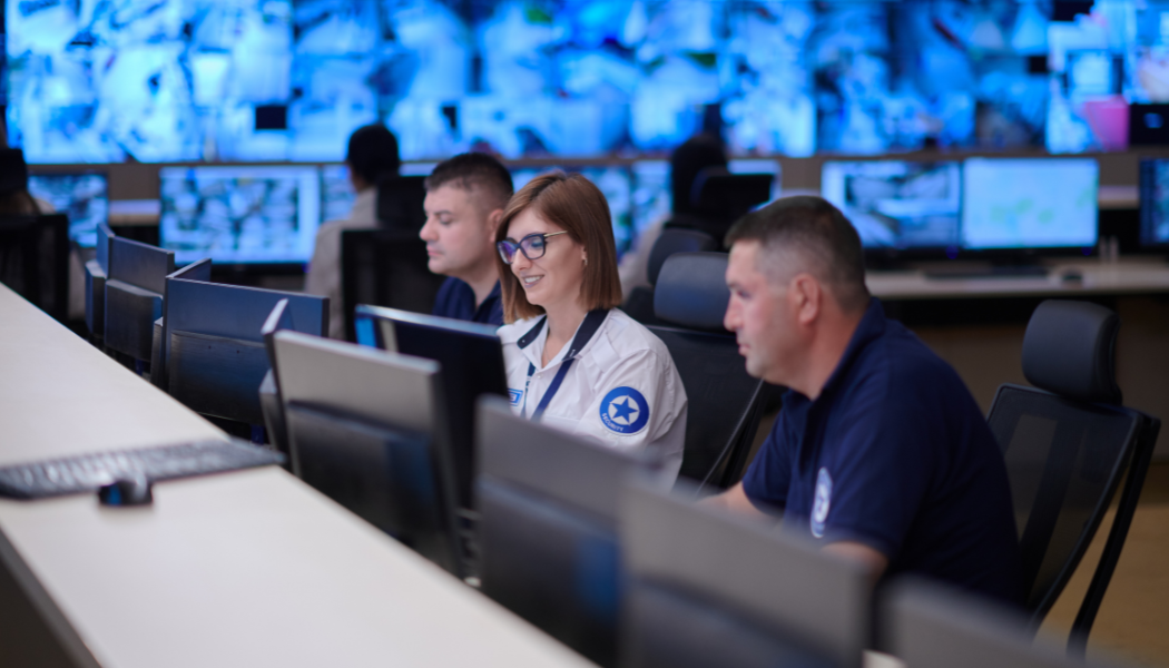 People working at computers in a control room with multiple screens and monitors.
