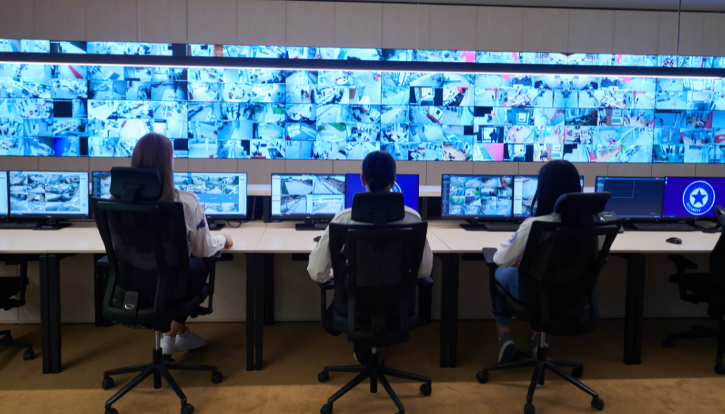 Three people sitting at desks with multiple monitors in a control room, monitoring a large wall of video feeds.