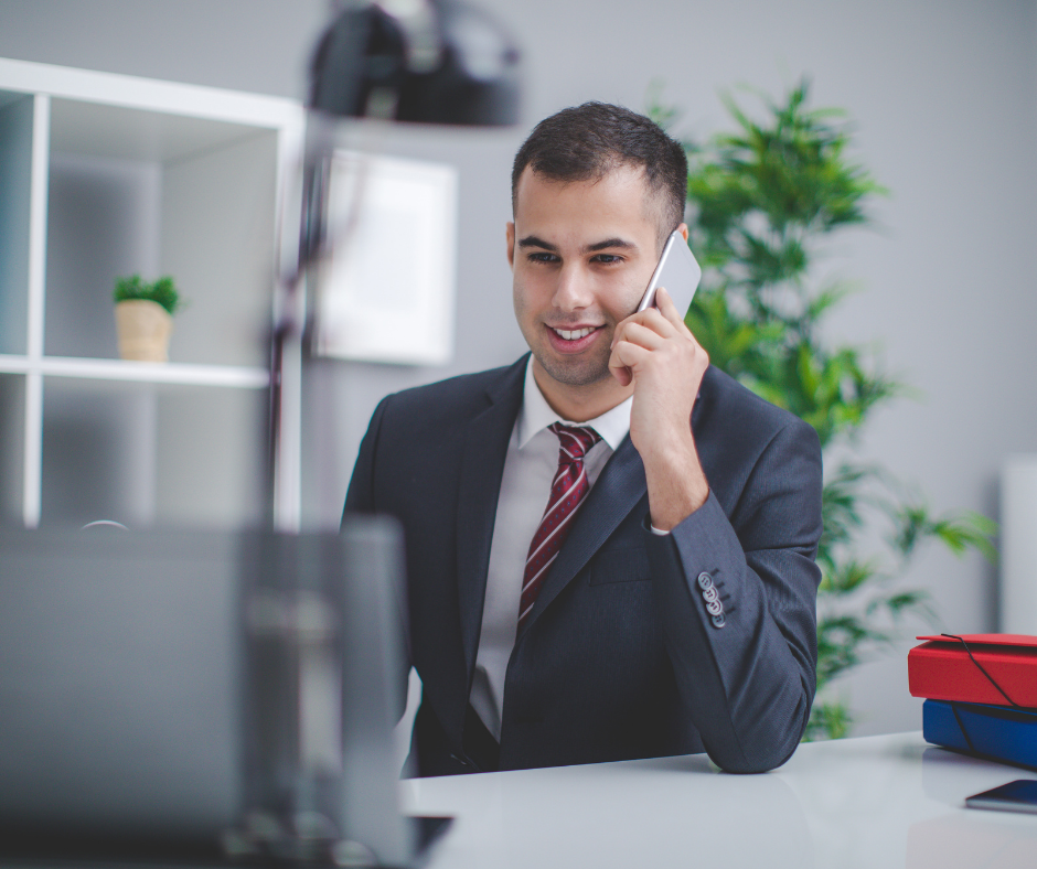 A young man in a business suit talking on a landline phone in an office setting, with a plant and shelves in the background.