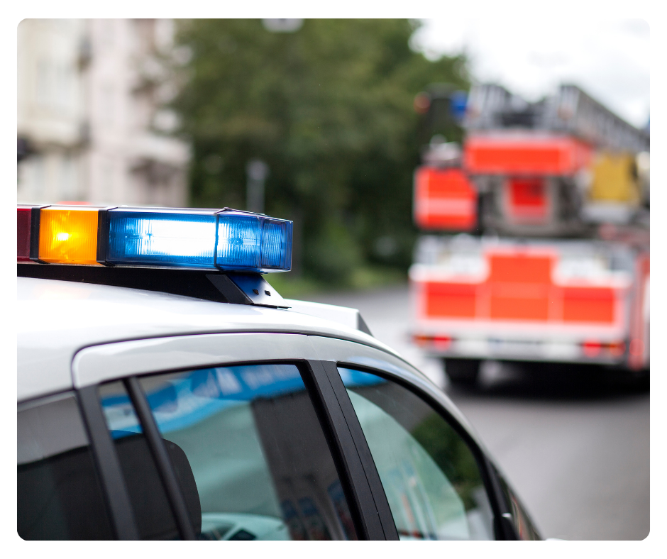 A police car with a flashing blue and yellow light on top, and a fire truck with orange and white markings in the background.