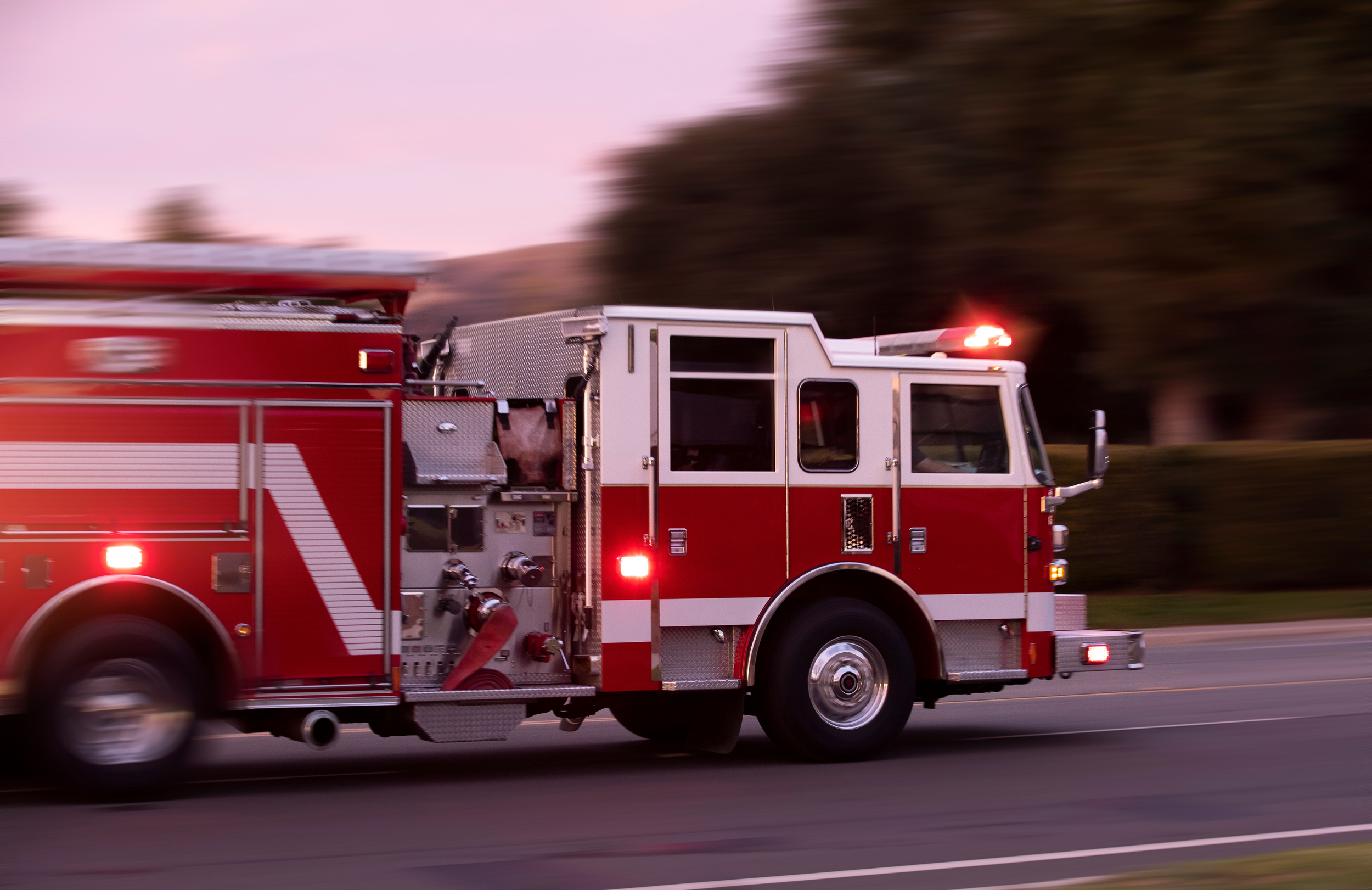 A red and white fire truck in motion on a road during sunset with blurred trees in the background.