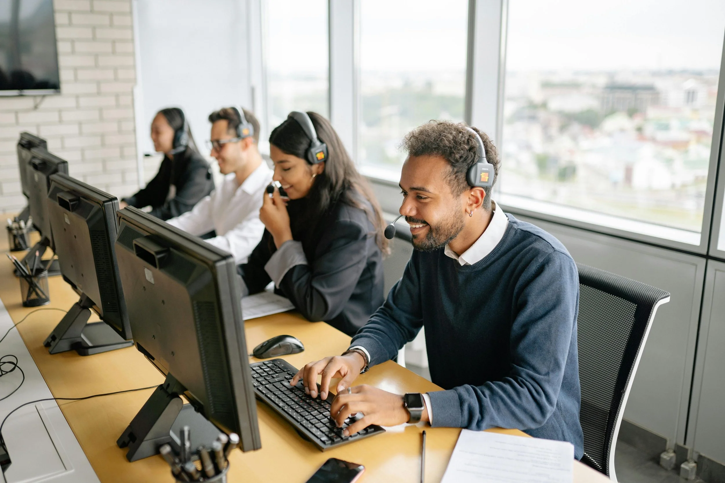 A diverse group of four customer service representatives working at desks in an office, wearing headsets and using computers, with large windows showing a city view in the background.