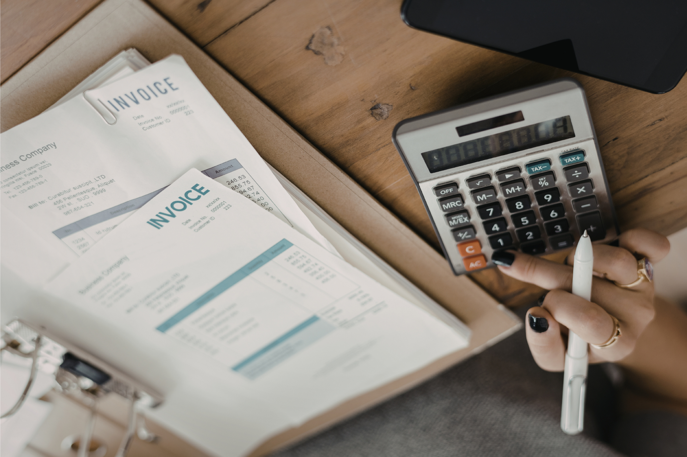 Hand using a calculator and pen beside stacked invoices on a wooden desk.