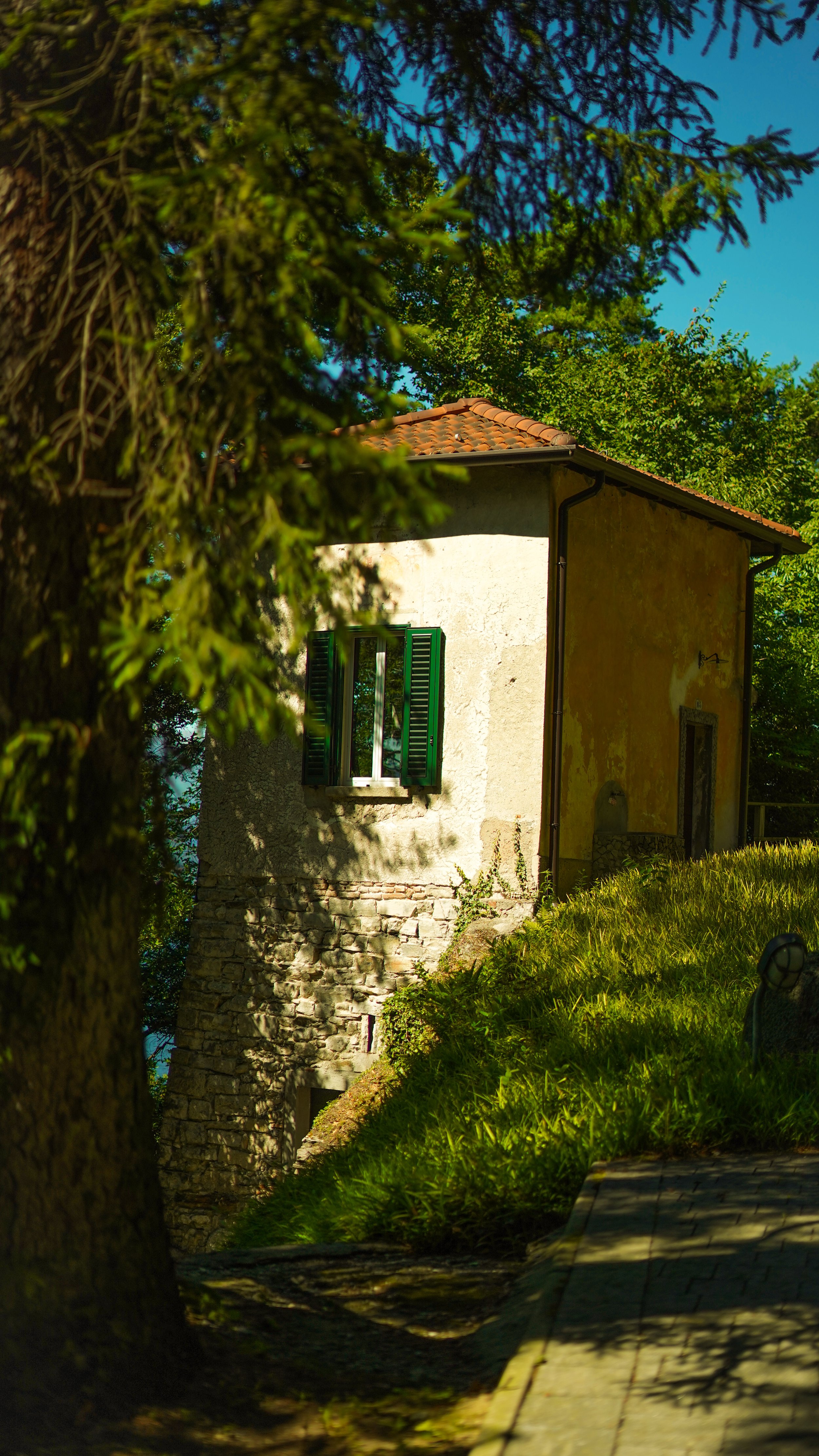 A small house with a tiled roof and open window shutters, surrounded by lush trees and greenery, with a cobblestone path nearby.