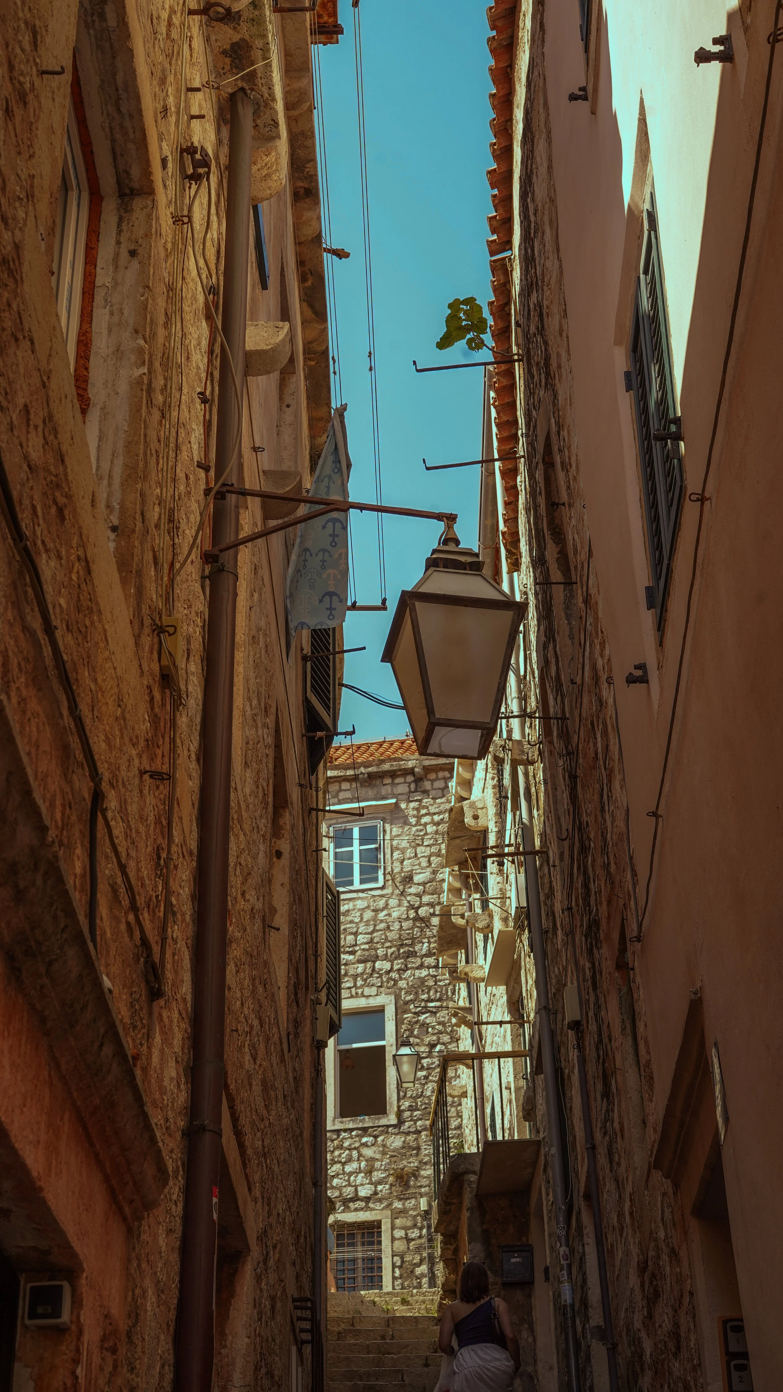 Narrow alleyway between old stone and stucco buildings with stairs at the end, a woman walking away, laundry hanging, and a street lamp.