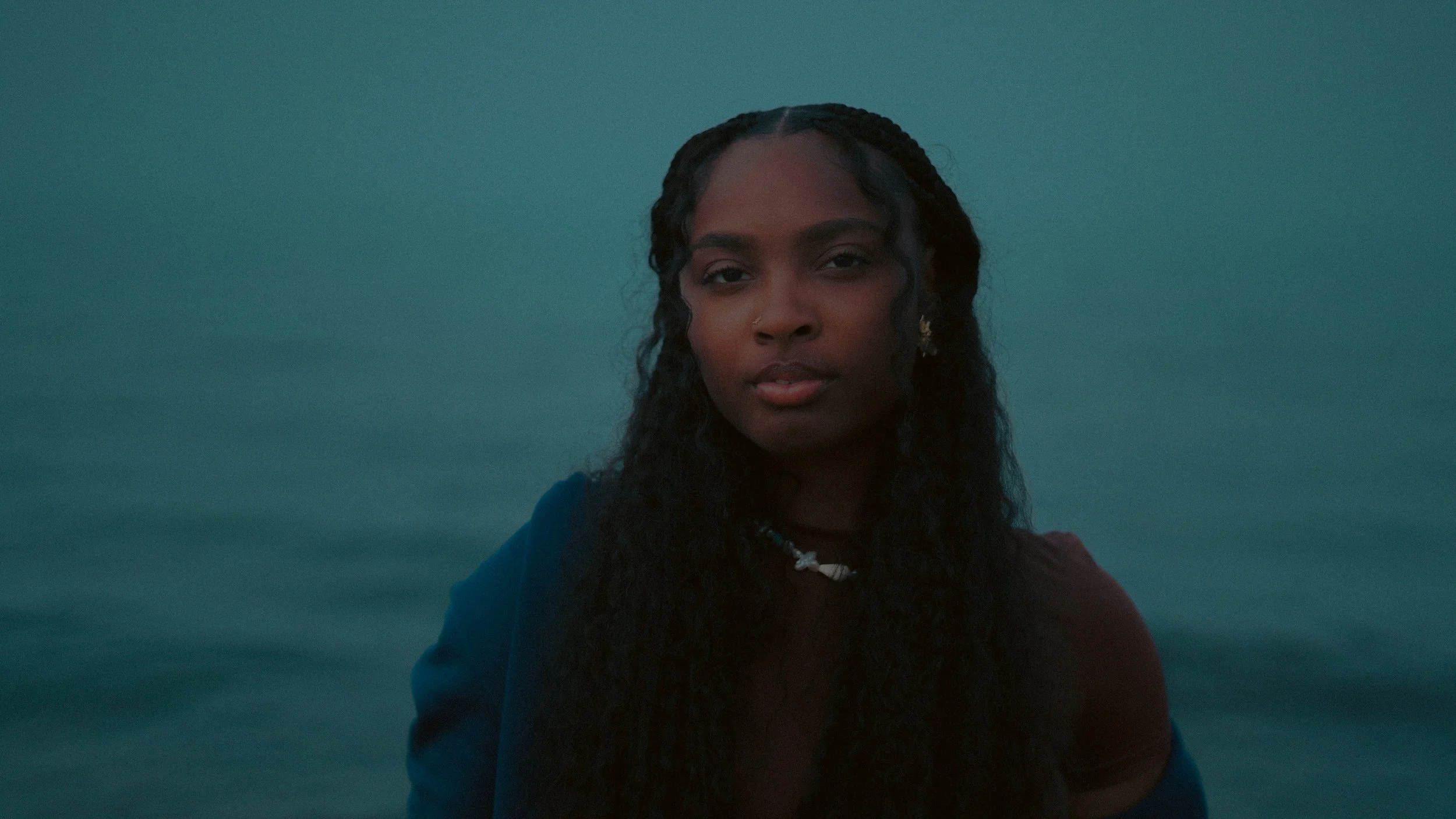 A young woman with long, curly black hair, wearing a navy jacket and a shell necklace, standing in front of a body of water with a calm, greenish-blue appearance during twilight.