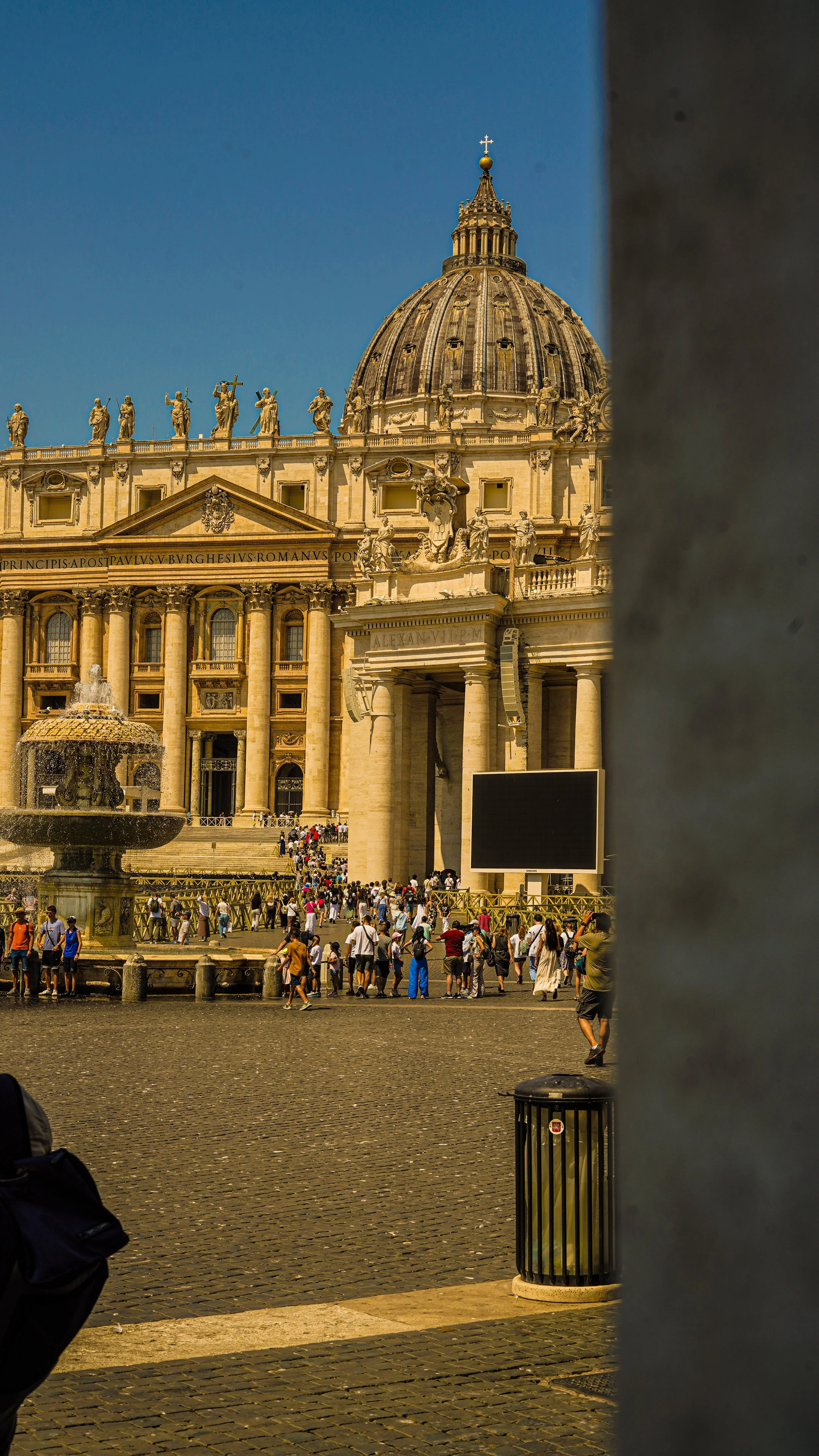 View of St. Peter's Basilica in Vatican City seen through a gap in a wall or fence, with a large crowd in the square in front.