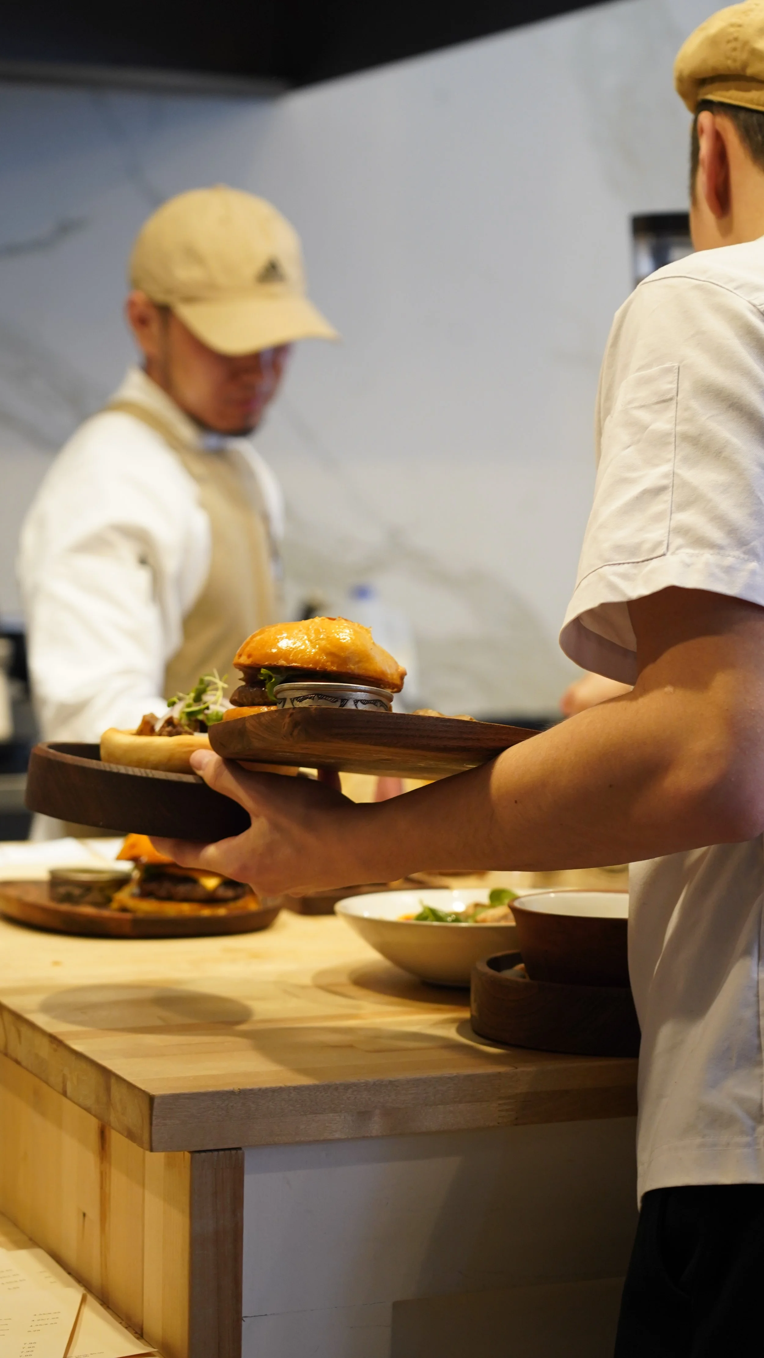 Person in a white shirt holding a wooden tray with a burger and other food items in a restaurant kitchen, with chefs in the background.