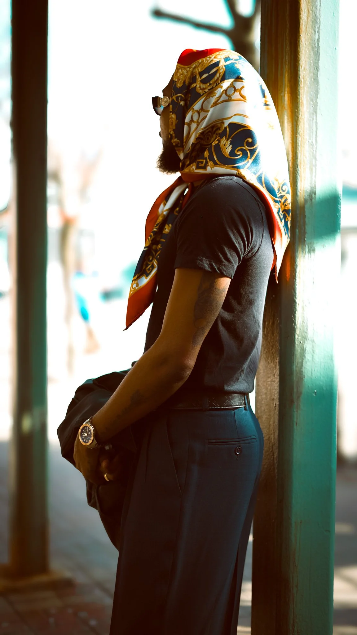 A man with a beard wearing a colorful scarf over his head, a black t-shirt, dark pants, and a watch, leaning against a support beam outside in natural light.