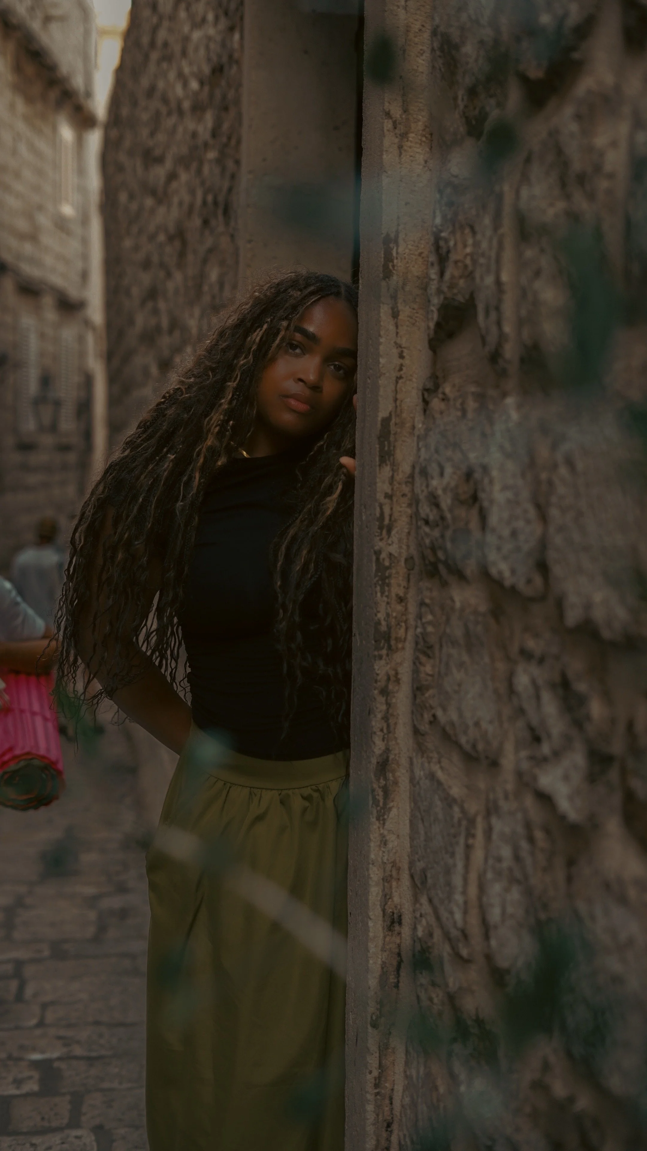 A woman with long curly hair peeking out from behind a stone wall in a narrow alleyway.
