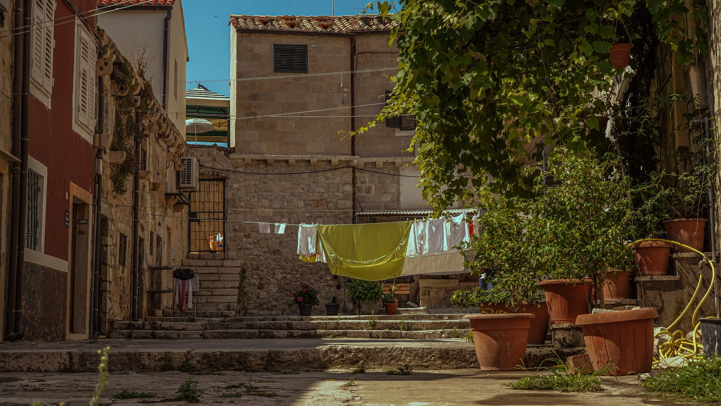 A cobblestone courtyard in an old European neighborhood with clothes hanging on a line to dry, potted plants along the staircase, and stone buildings with shutters and air conditioning units.