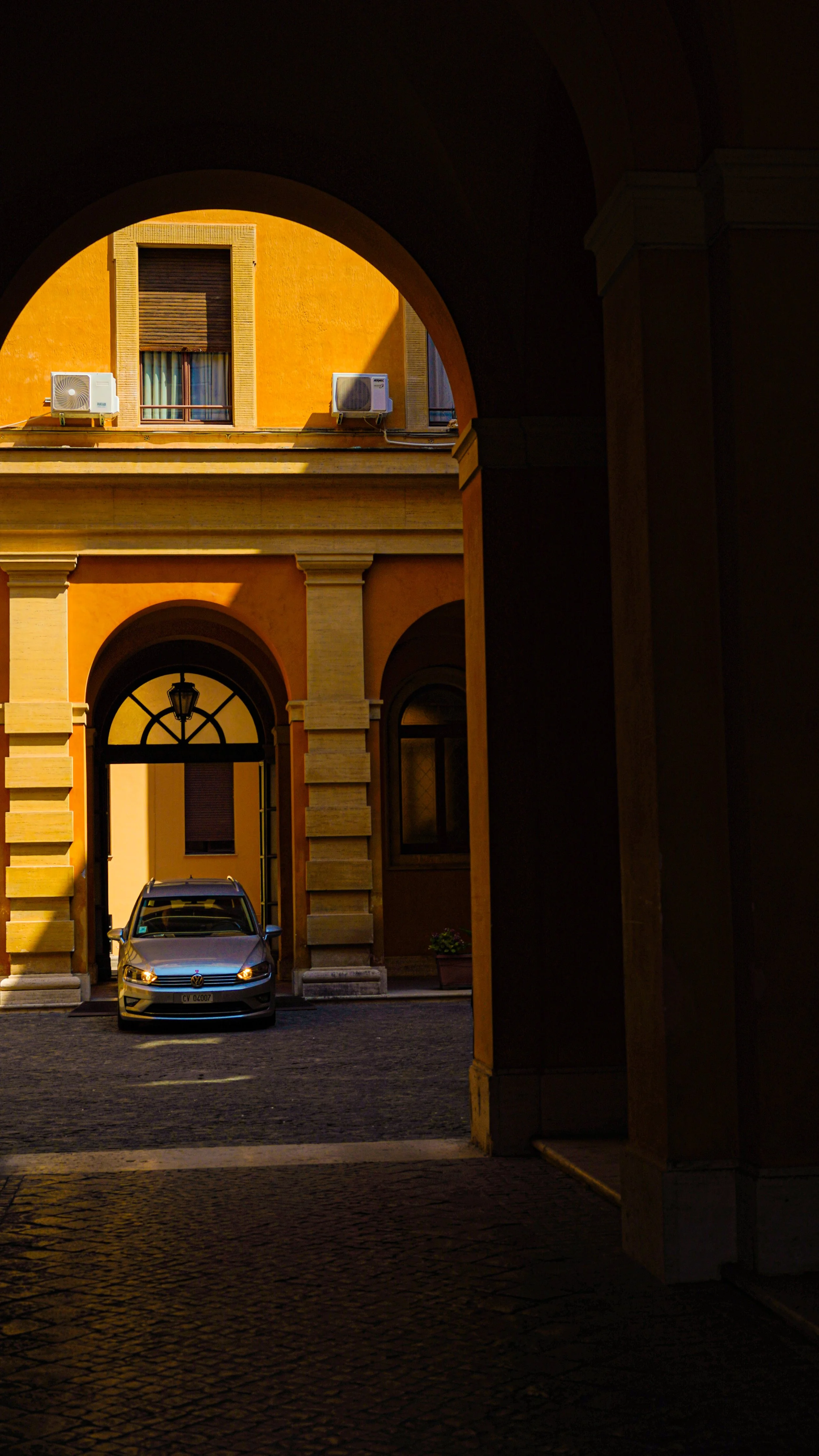 A silver Volkswagen car parked under an archway in front of a yellow building with windows and air conditioning units, during sunset casting shadows.