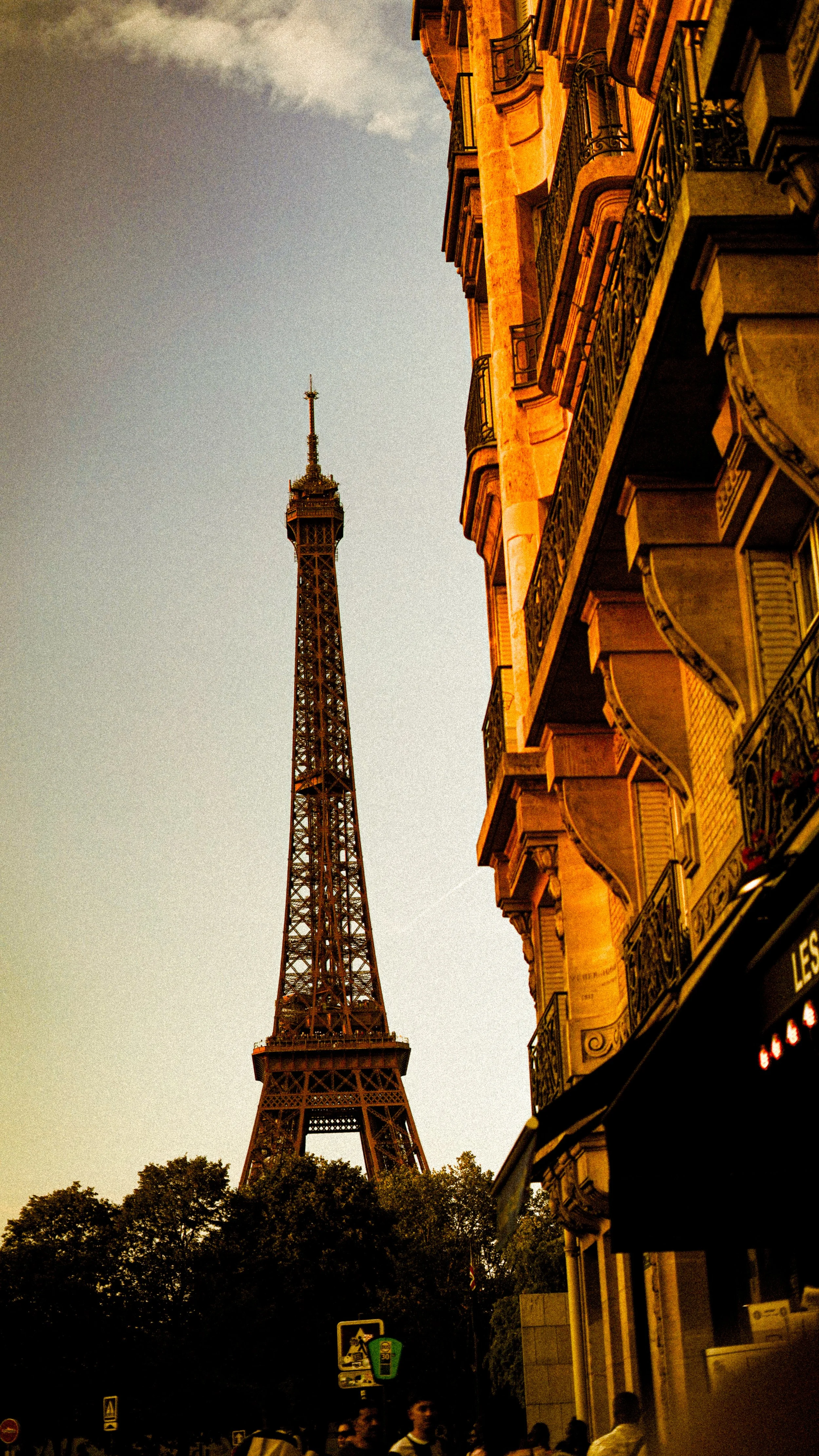 The Eiffel Tower in Paris, seen from a street with buildings and people below, during dusk or early evening.