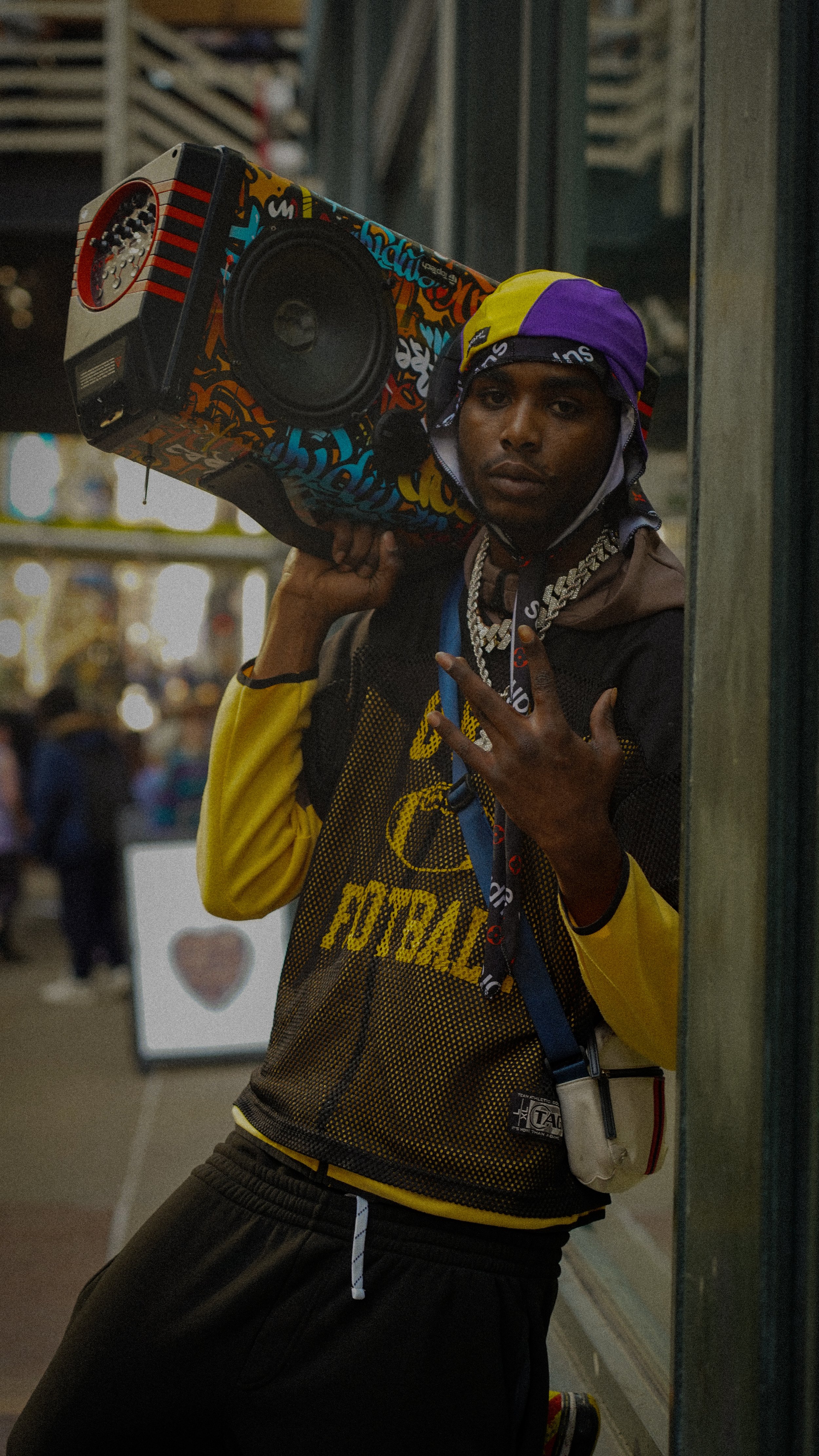 A young man with dark skin wearing a purple and yellow bandana, a chain necklace, and a black and yellow football jersey, holding a colorful speaker on his shoulder in a lively, crowded street or market.