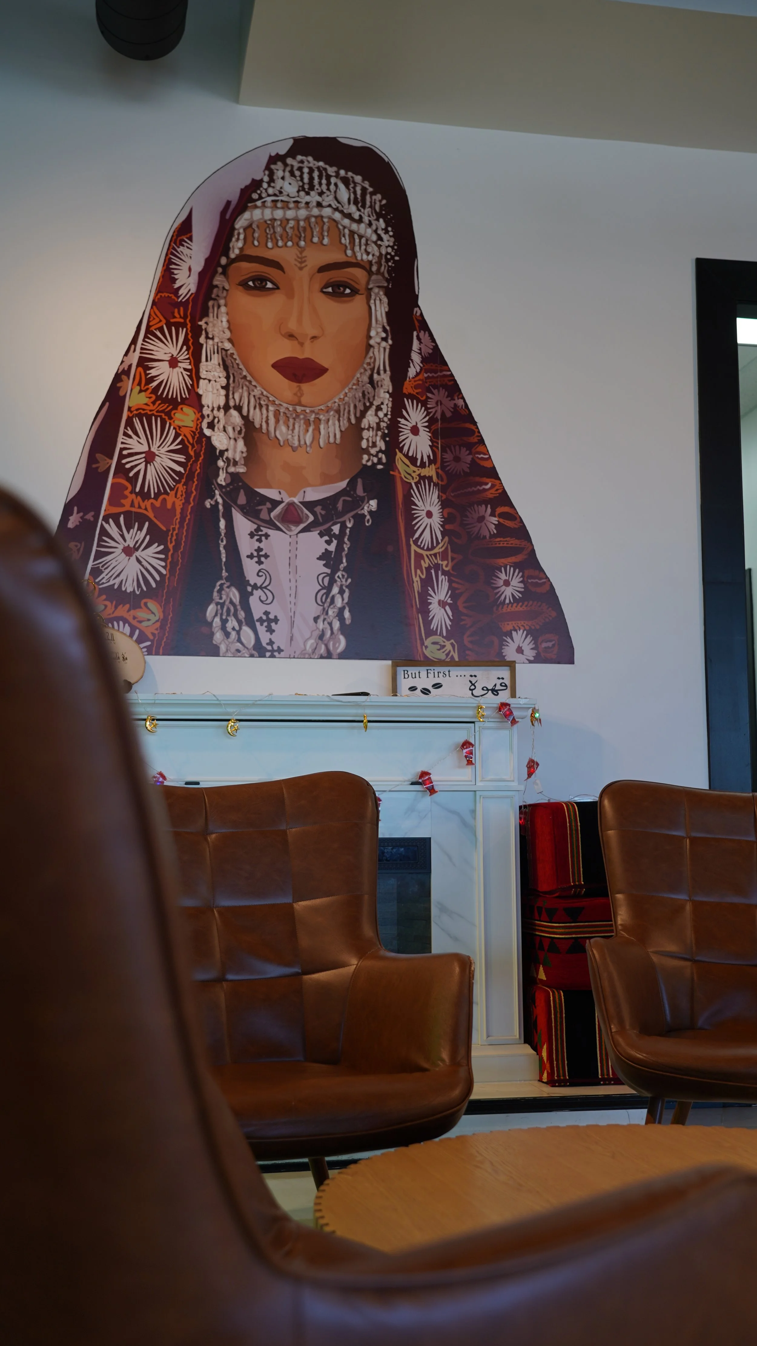 Portrait of a woman in traditional dress with jewelry and a headscarf, displayed on a large wall poster in a room with brown leather chairs and a white fireplace.