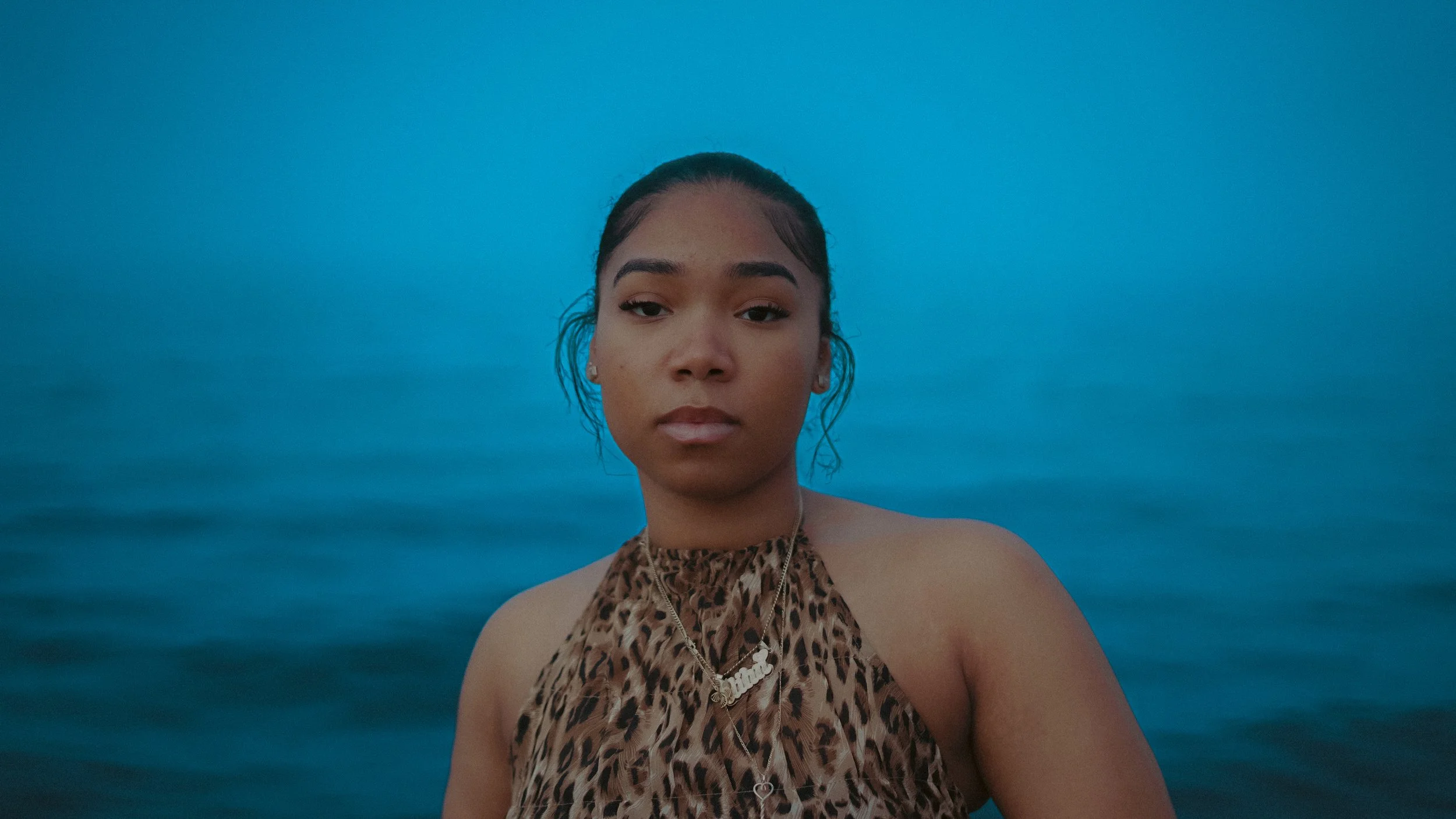 A woman wearing a leopard print top and layered necklaces takes a selfie in front of a calm body of water with a clear blue sky overhead.