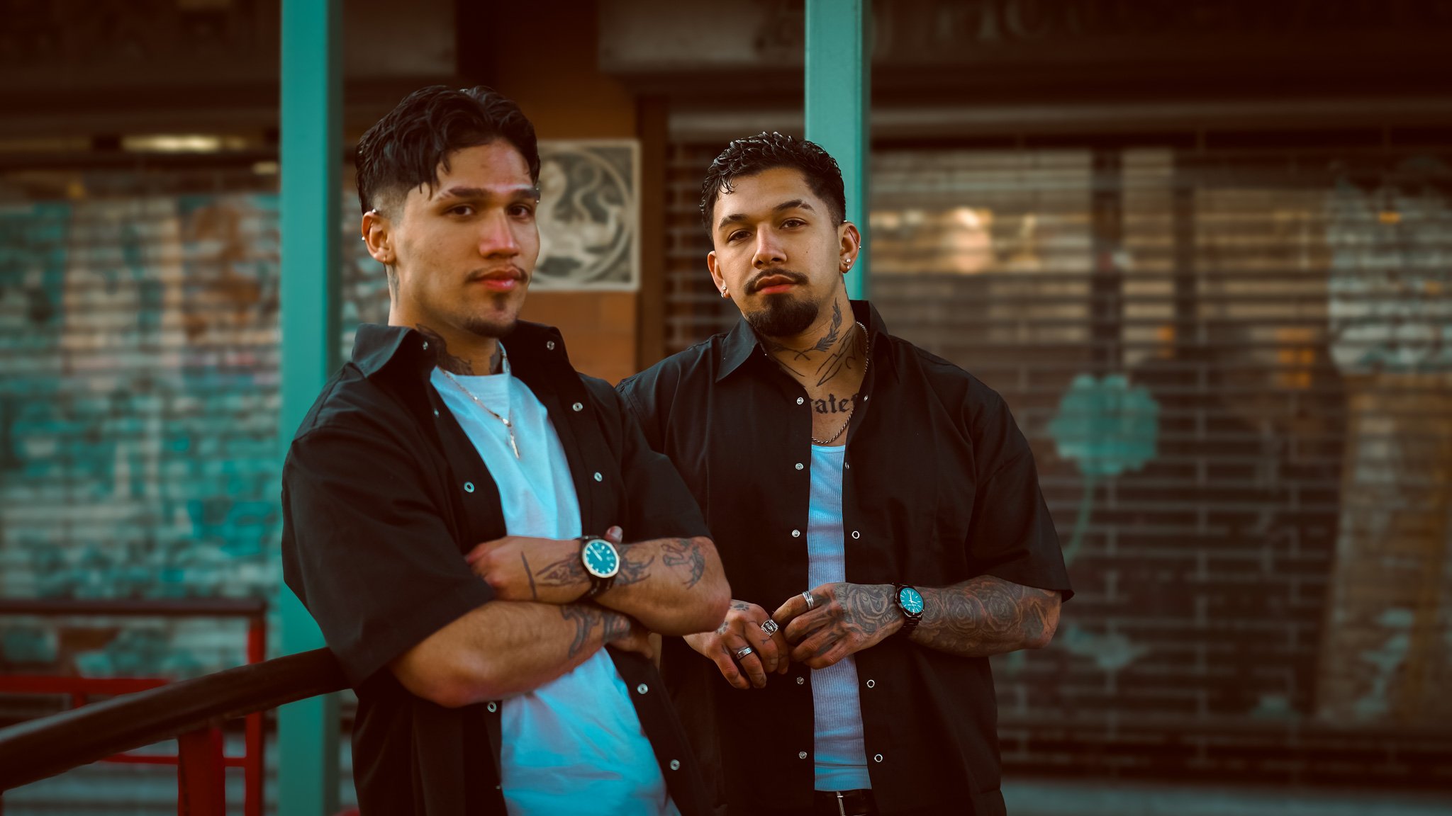 Two young men with tattoos, wearing black open shirts over white T-shirts, standing outdoors and looking at the camera with serious expressions.