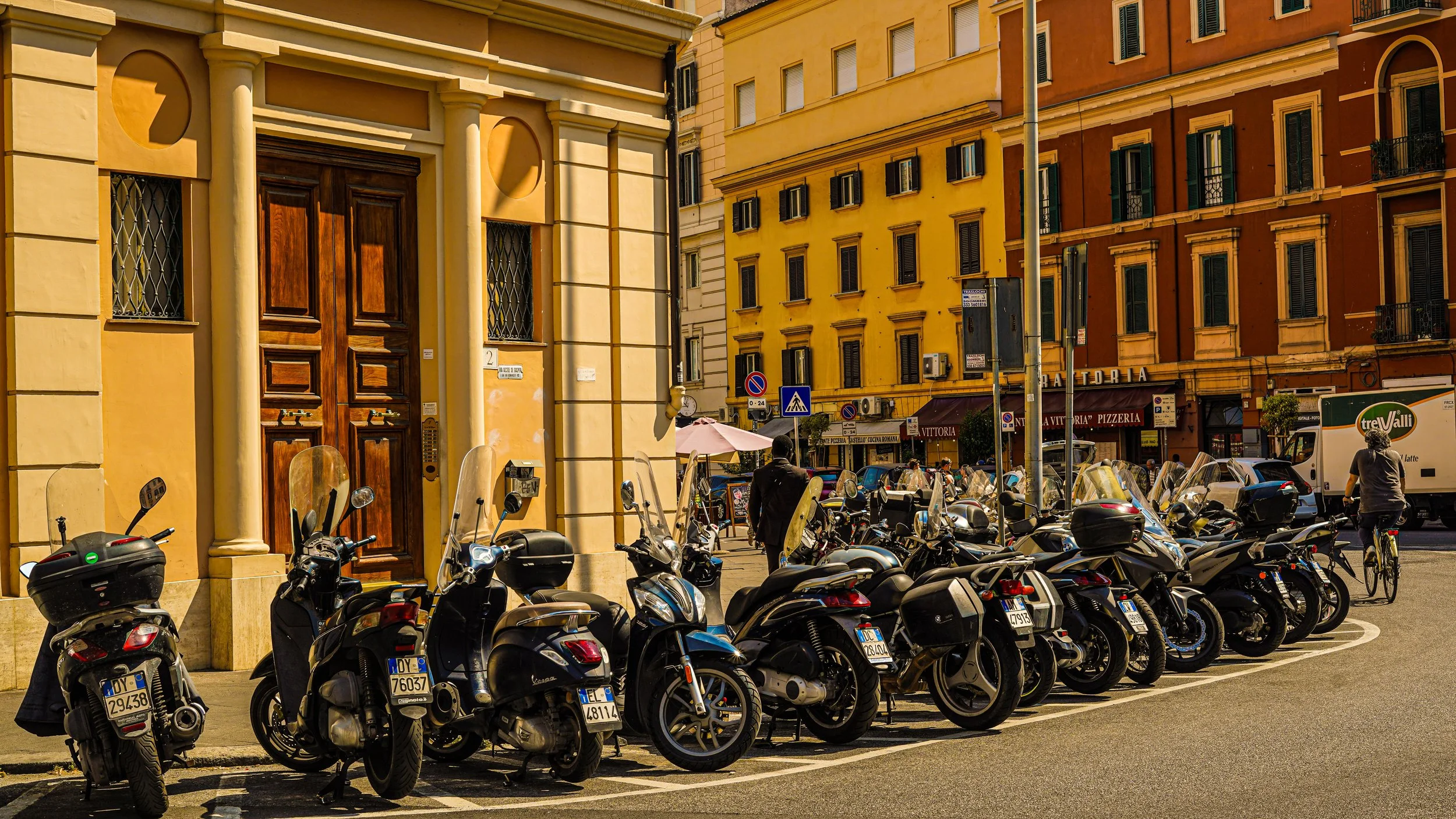 Motorcycles parked along a city street in front of a yellow building with a large wooden door. People are walking and cycling nearby, with colorful buildings and shops in the background.