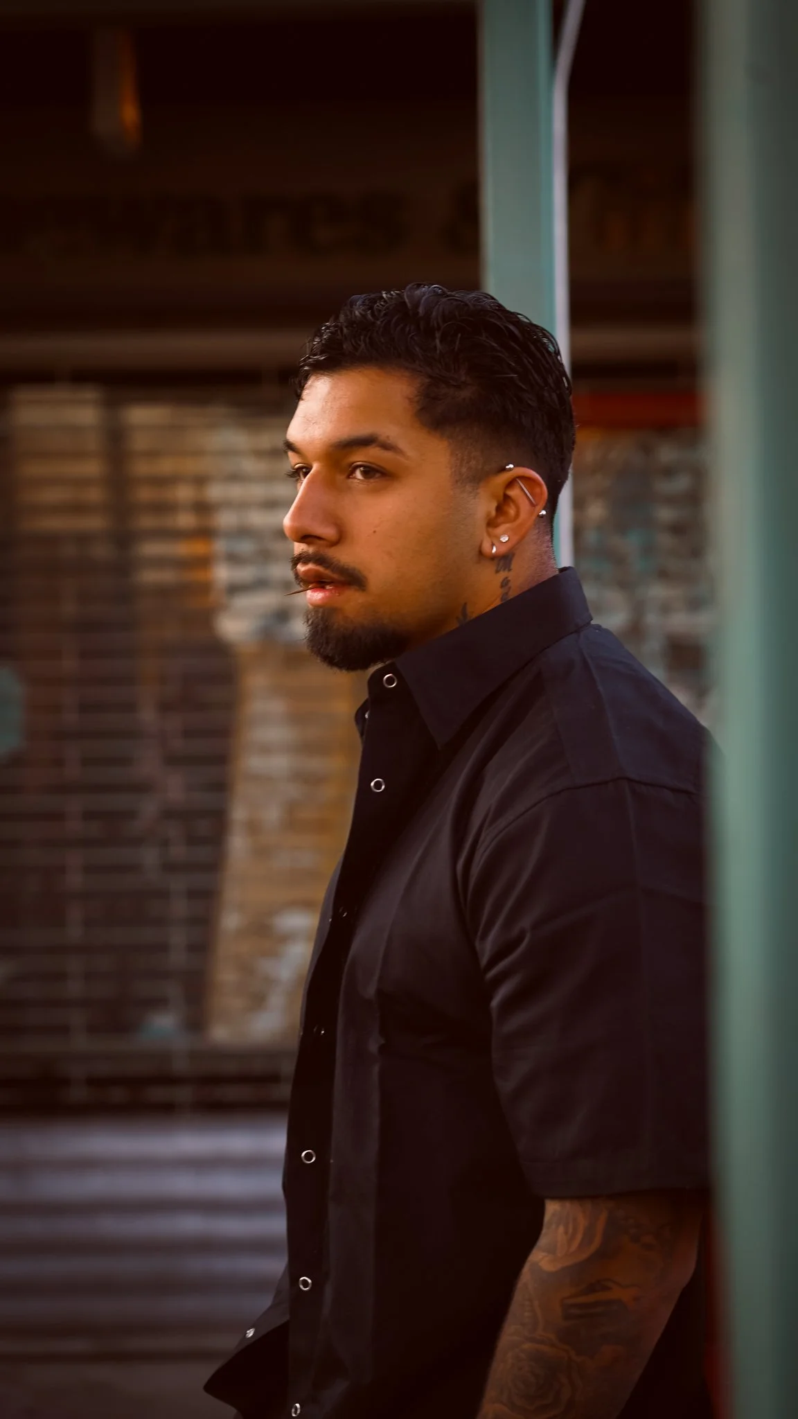 A young man with dark, styled hair, facial hair, multiple earrings, and tattoos on his neck and arm, wearing a black shirt, standing against a brick wall and looking to the side.
