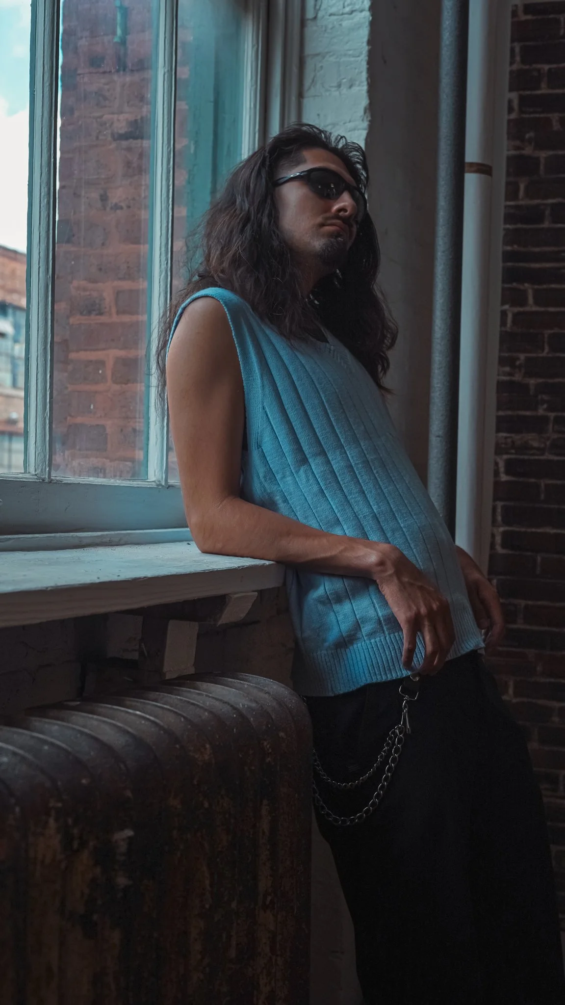 Person with long dark hair wearing sunglasses and a sleeveless blue shirt, leaning against a windowsill in an industrial-style room with brick walls.