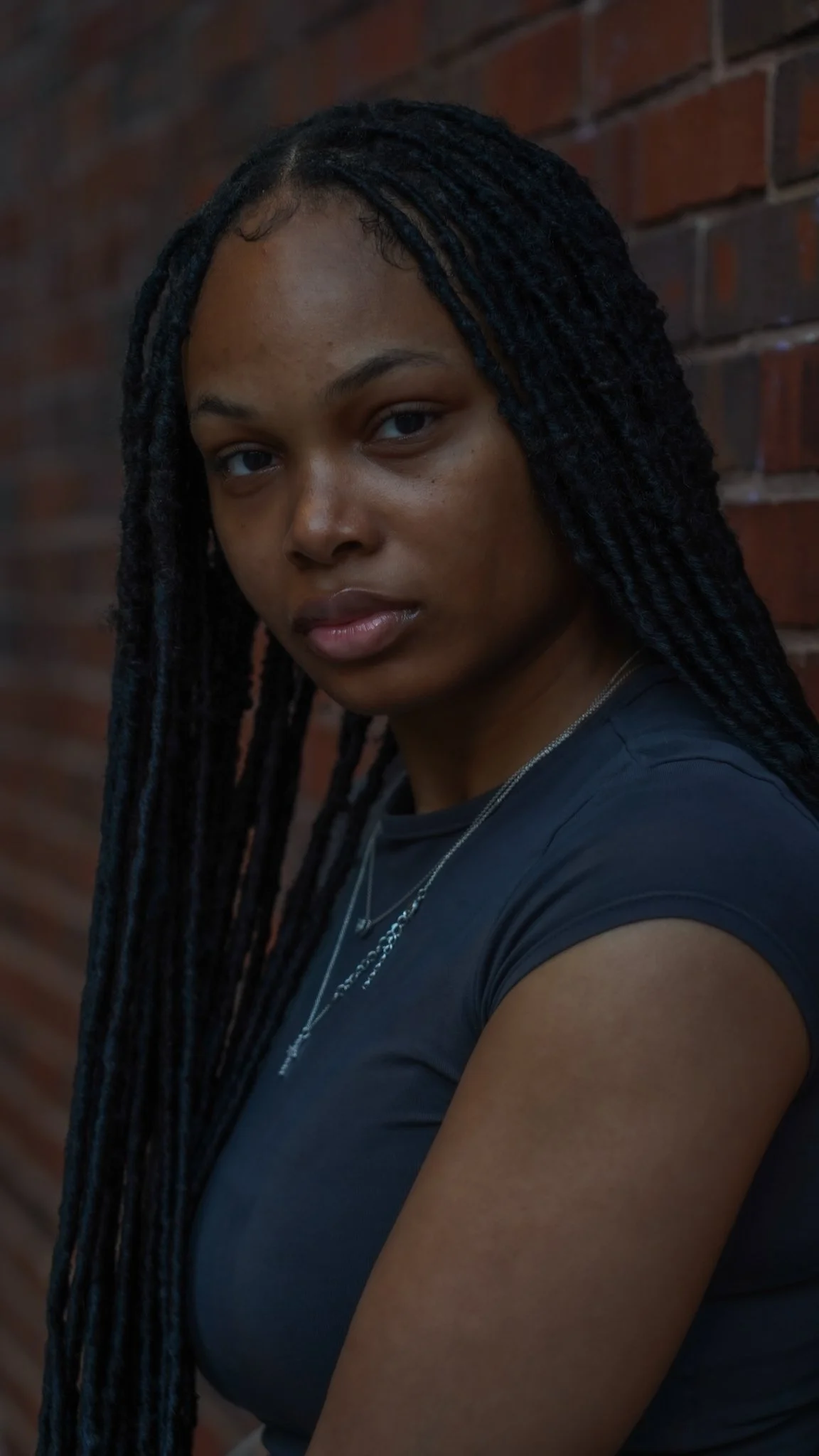 A young woman with long braids and a black shirt standing against a brick wall, looking at the camera.