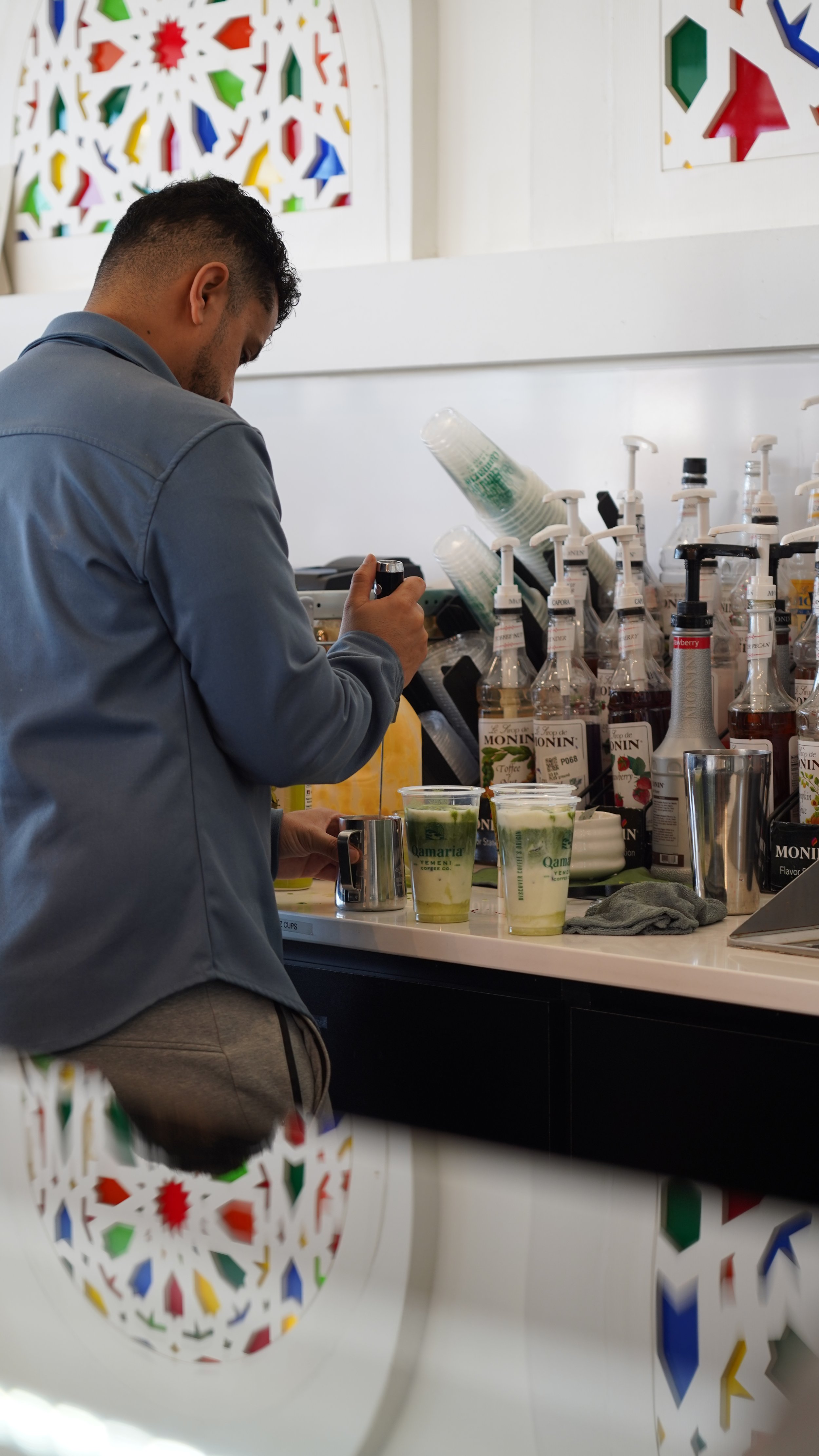 A person preparing drinks at a counter with various bottles of syrups and cups, two cups filled with a green and white drink, with colorful stained glass window panels in the background.