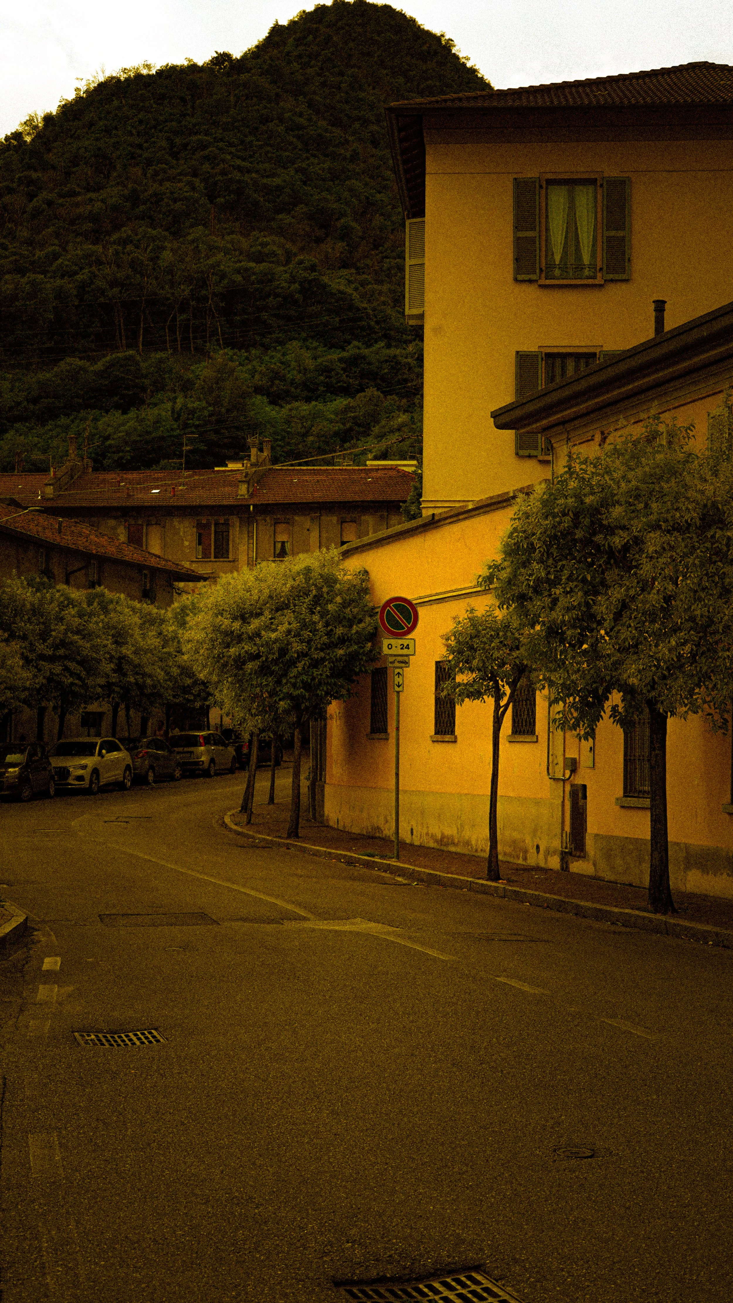 Empty street in a European town with parked cars, trees, a no parking sign, and a yellow building with green window shutters, mountains in the background during dusk.