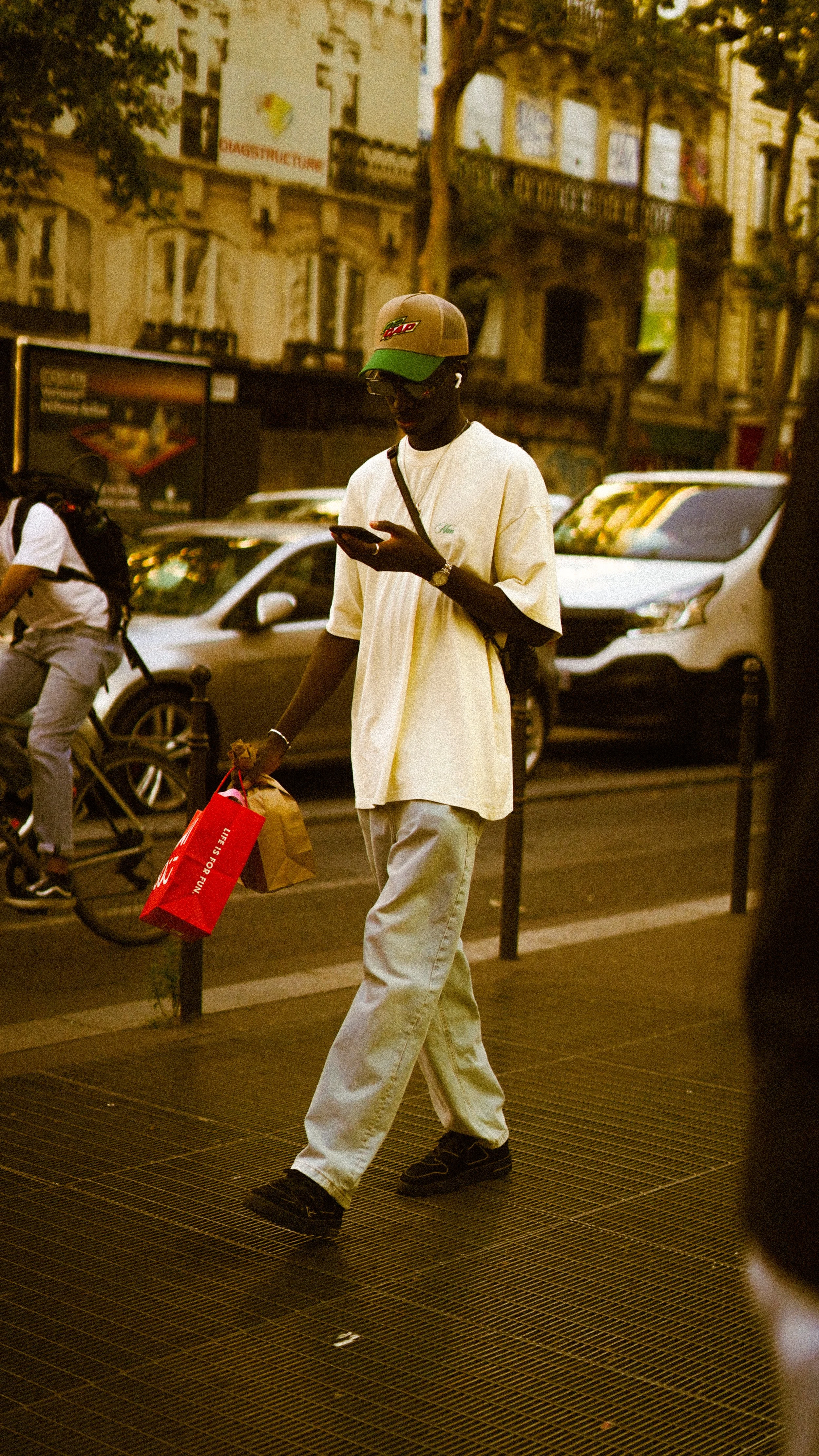 A man wearing a beige cap, sunglasses, a loose white t-shirt, and light-colored jeans walking on a city street, carrying a paper bag and a red shopping bag, while looking at his phone.