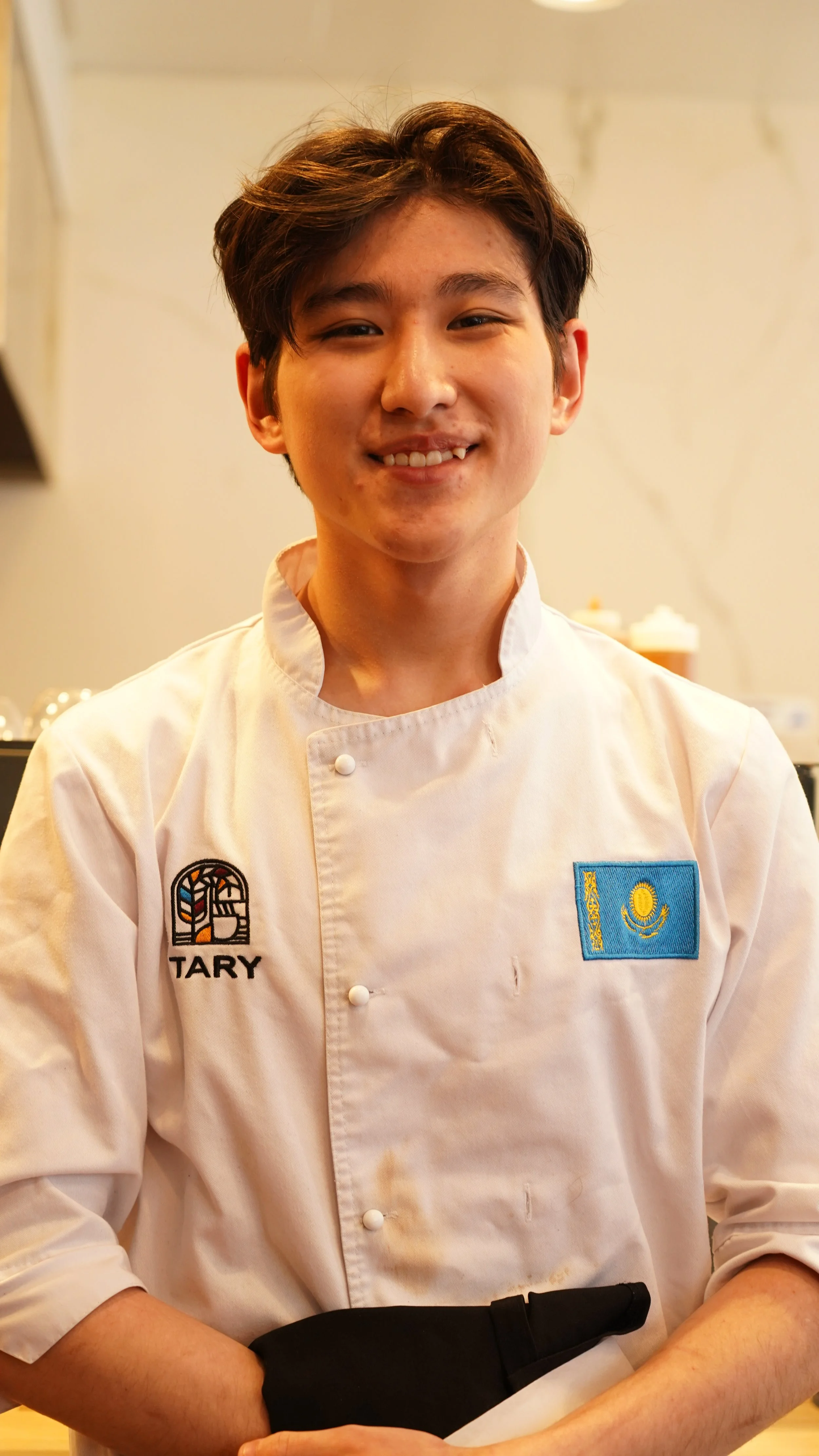 A young man in a white chef's coat with a Kazakh flag patch and a badge with an emblem, smiling, standing in a kitchen or food preparation area.