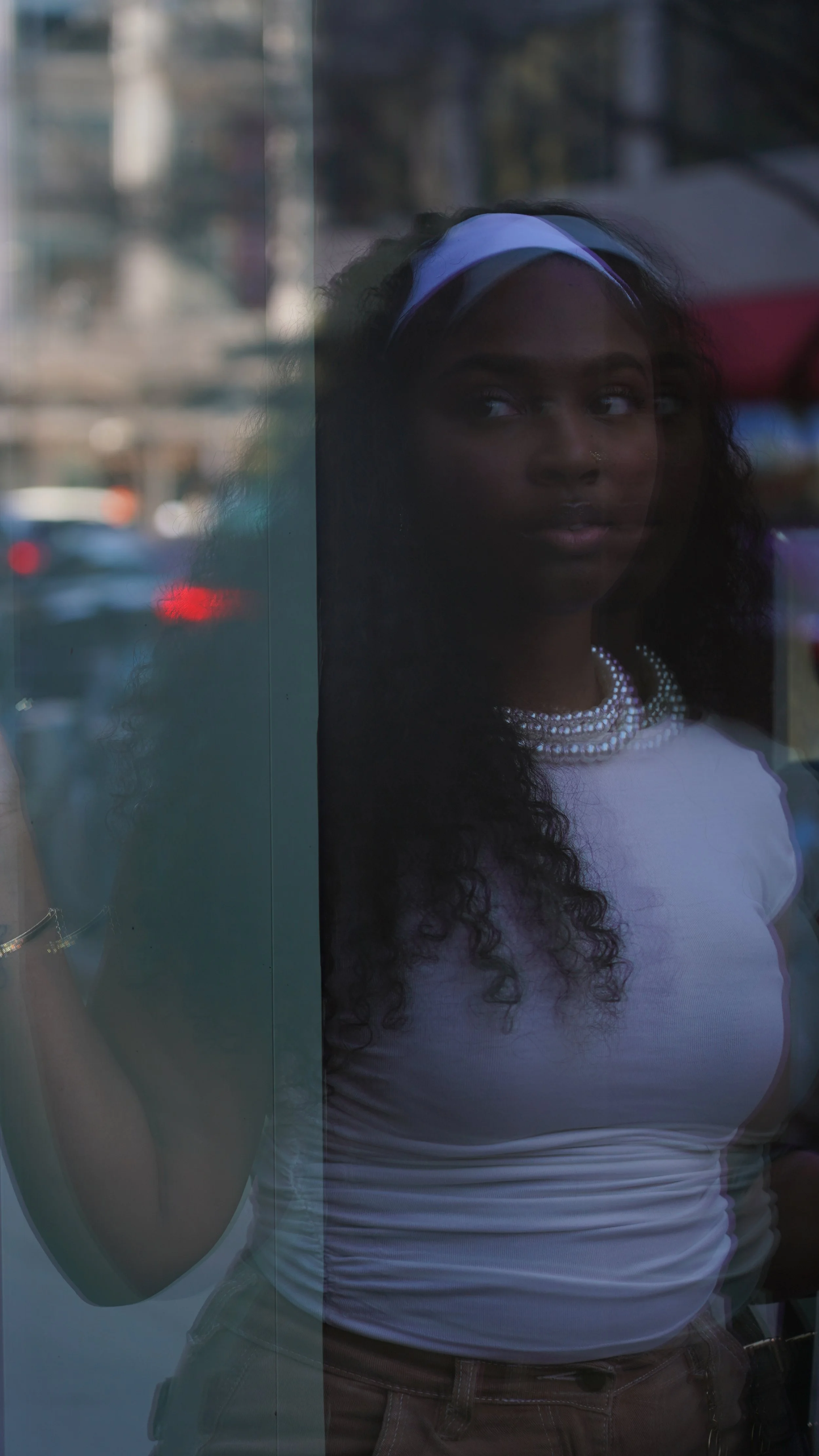 A woman with dark, curly hair and a bone-colored top, wearing a beaded necklace and a white headband, looks outside through a glass window that reflects the cityscape behind her.