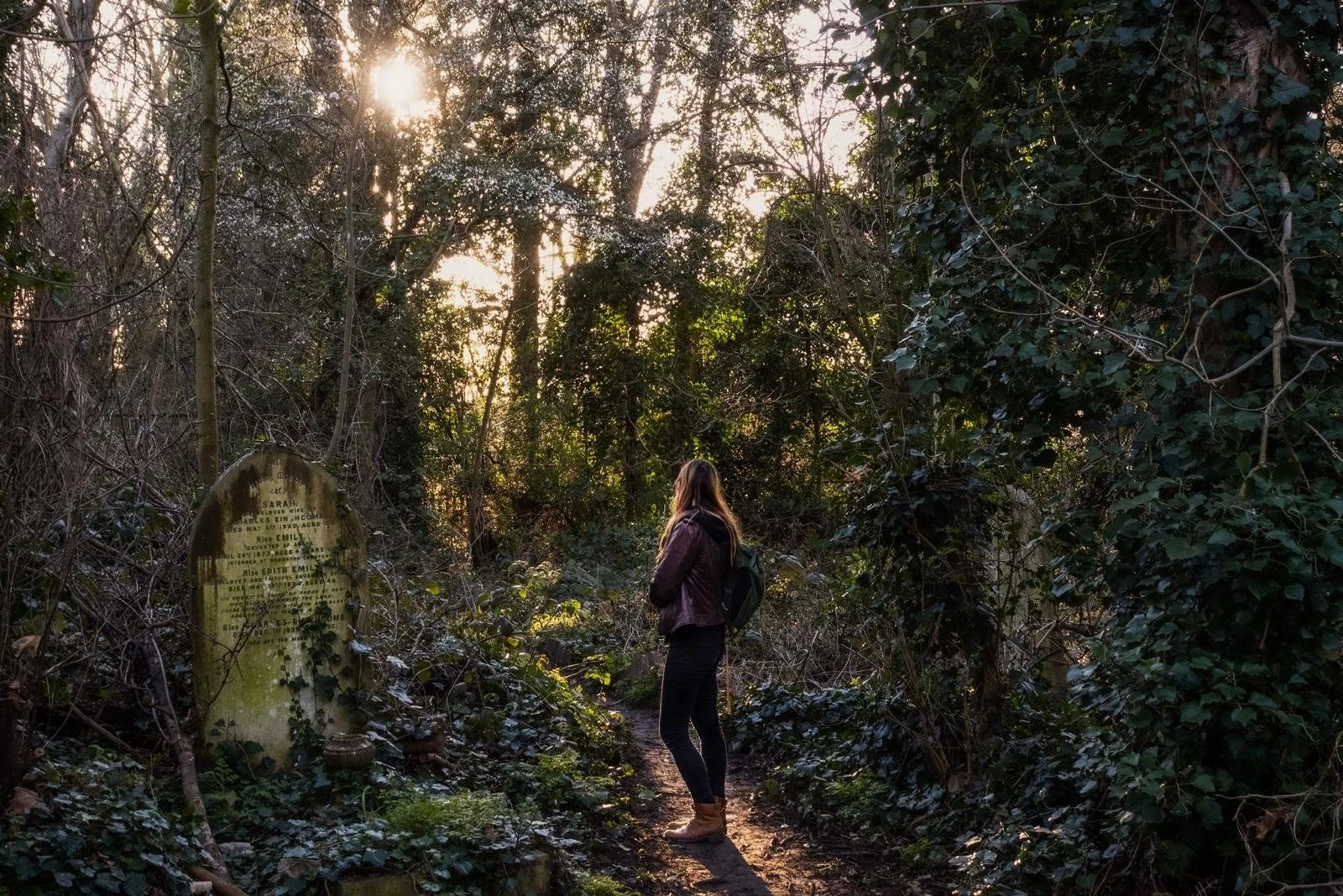 A woman standing on a forest trail during sunset, surrounded by dense trees and bushes, with a weathered historic tombstone on her left.