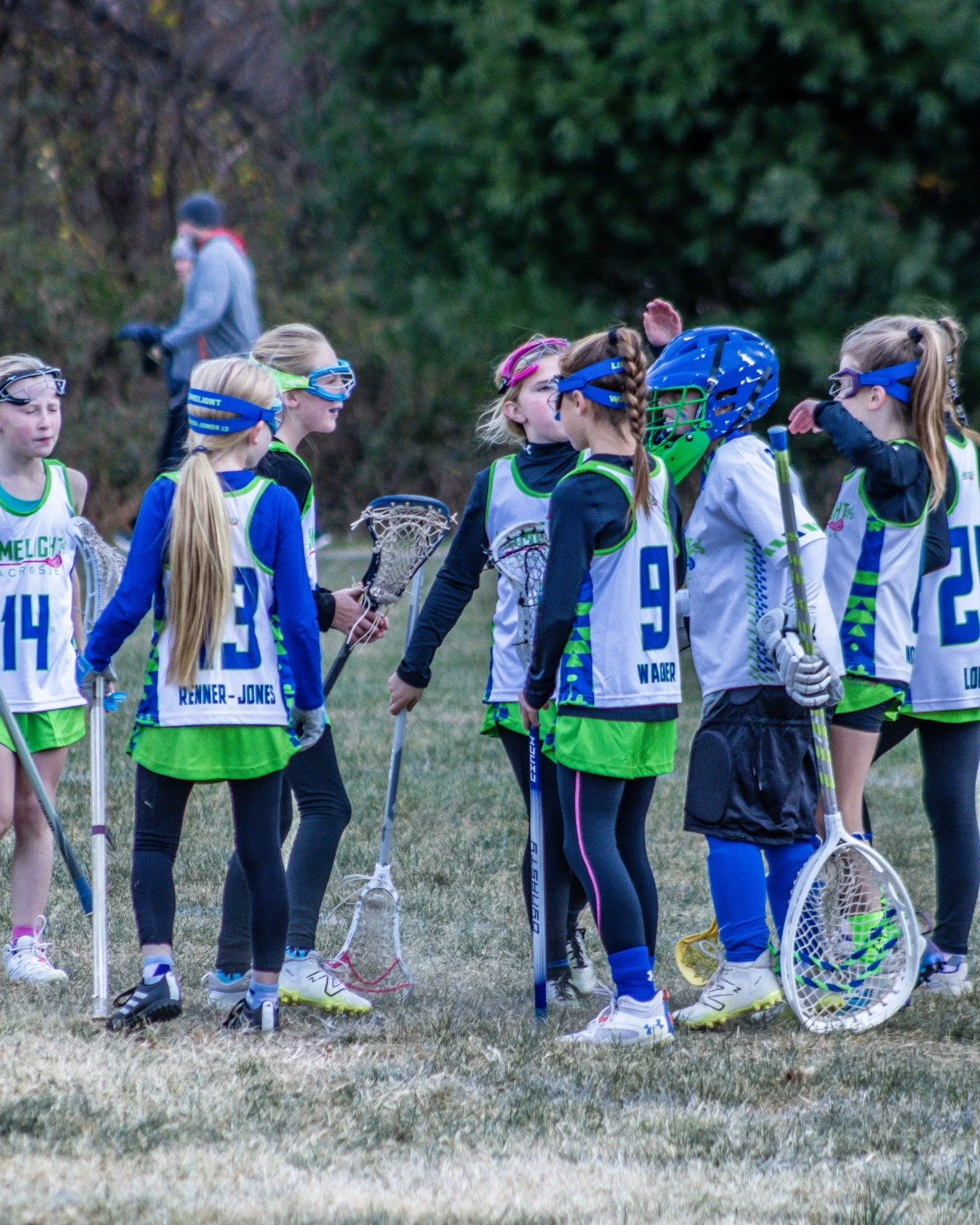 Sports photography Baltimore Maryland DMV. Girls lacrosse team huddling on a field, wearing uniforms and protective gear, some holding lacrosse sticks, in an outdoor setting with trees in the background.