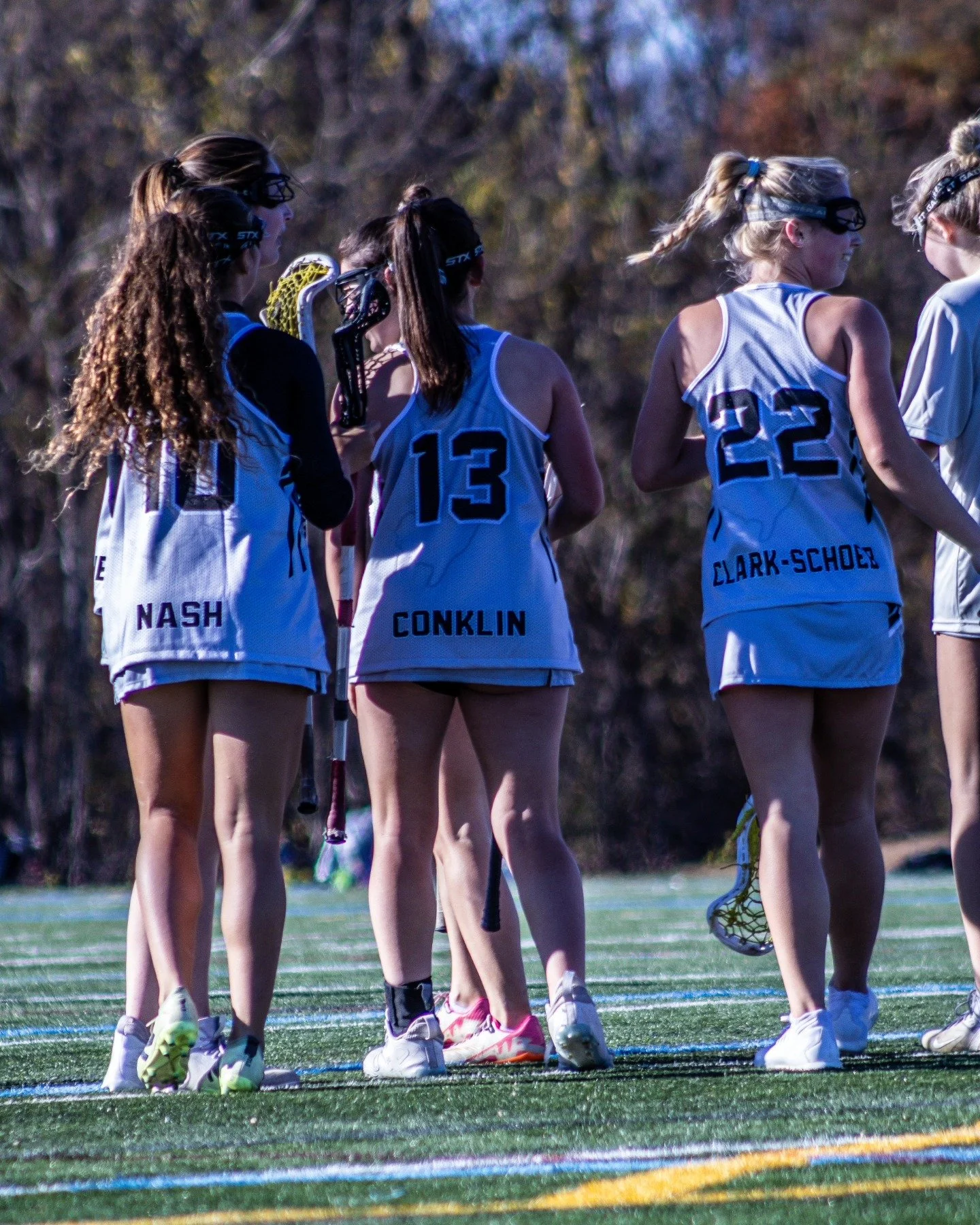 Sports photography Baltimore Maryland DMV.Female field hockey players standing on a field huddled together, wearing uniforms with numbers and names, holding field hockey sticks, some with protective goggles, possibly preparing for a game or practice.