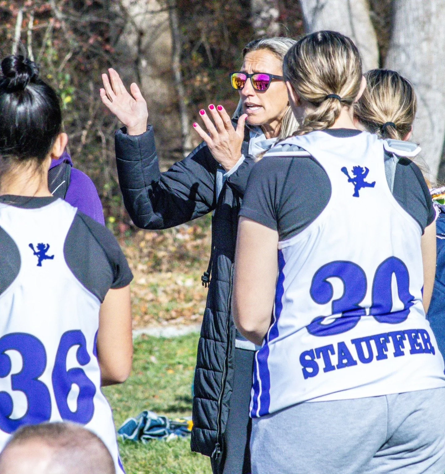 Sports photography Baltimore Maryland DMV. A female coach giving instructions or encouragement to young female soccer players during a game outdoors, with trees and fallen leaves in the background.