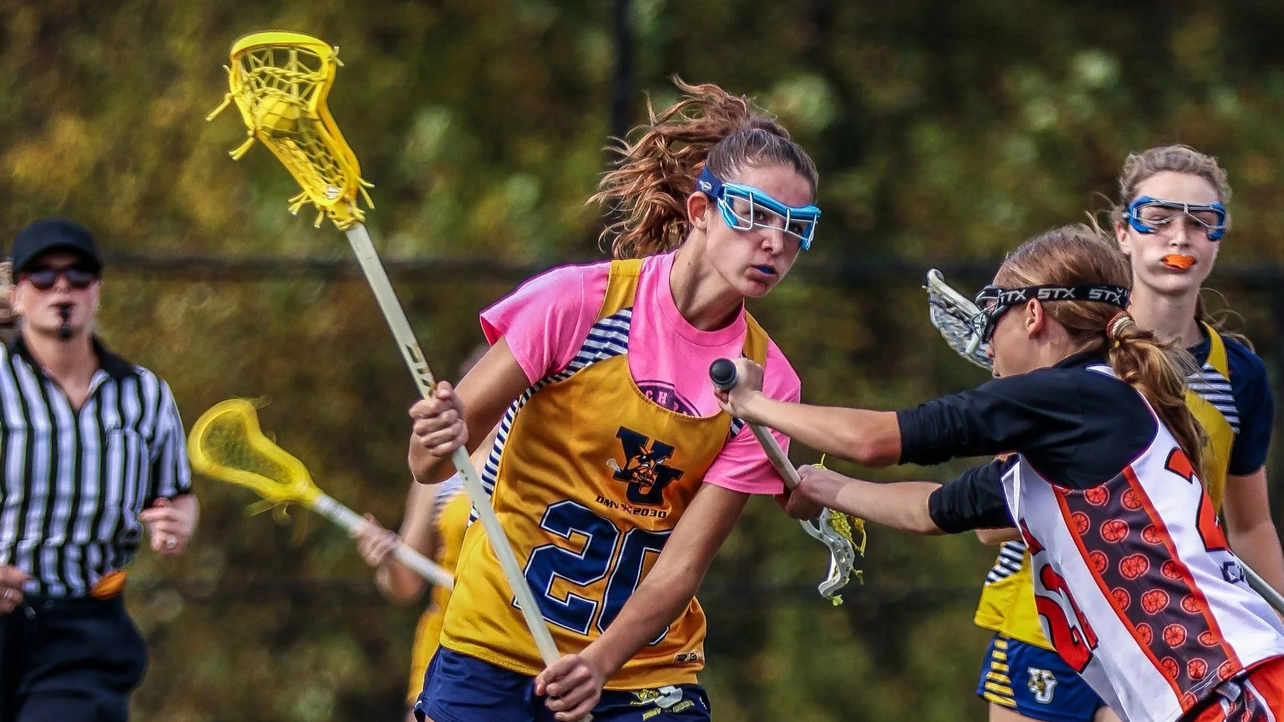 Sports photography Baltimore Maryland DMV. Lacrosse players in action, with one female player in a yellow jersey and pink shirt, holding a lacrosse stick, and others around her during a game outdoors.