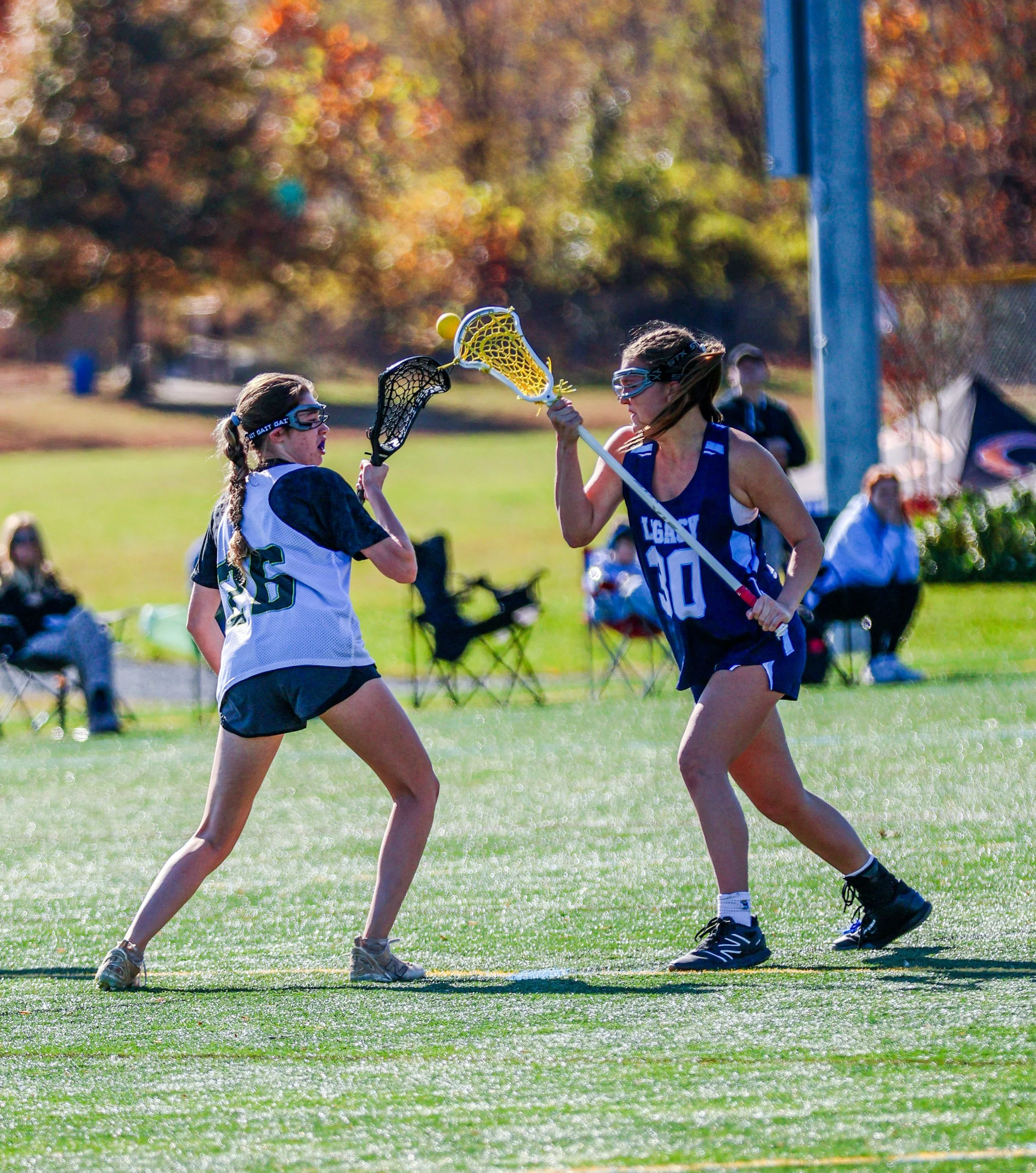 Sports photography Baltimore Maryland DMV. Two female lacrosse players compete on a sunny field, one with a white jersey and the other with a blue jersey, while several spectators watch in the background.