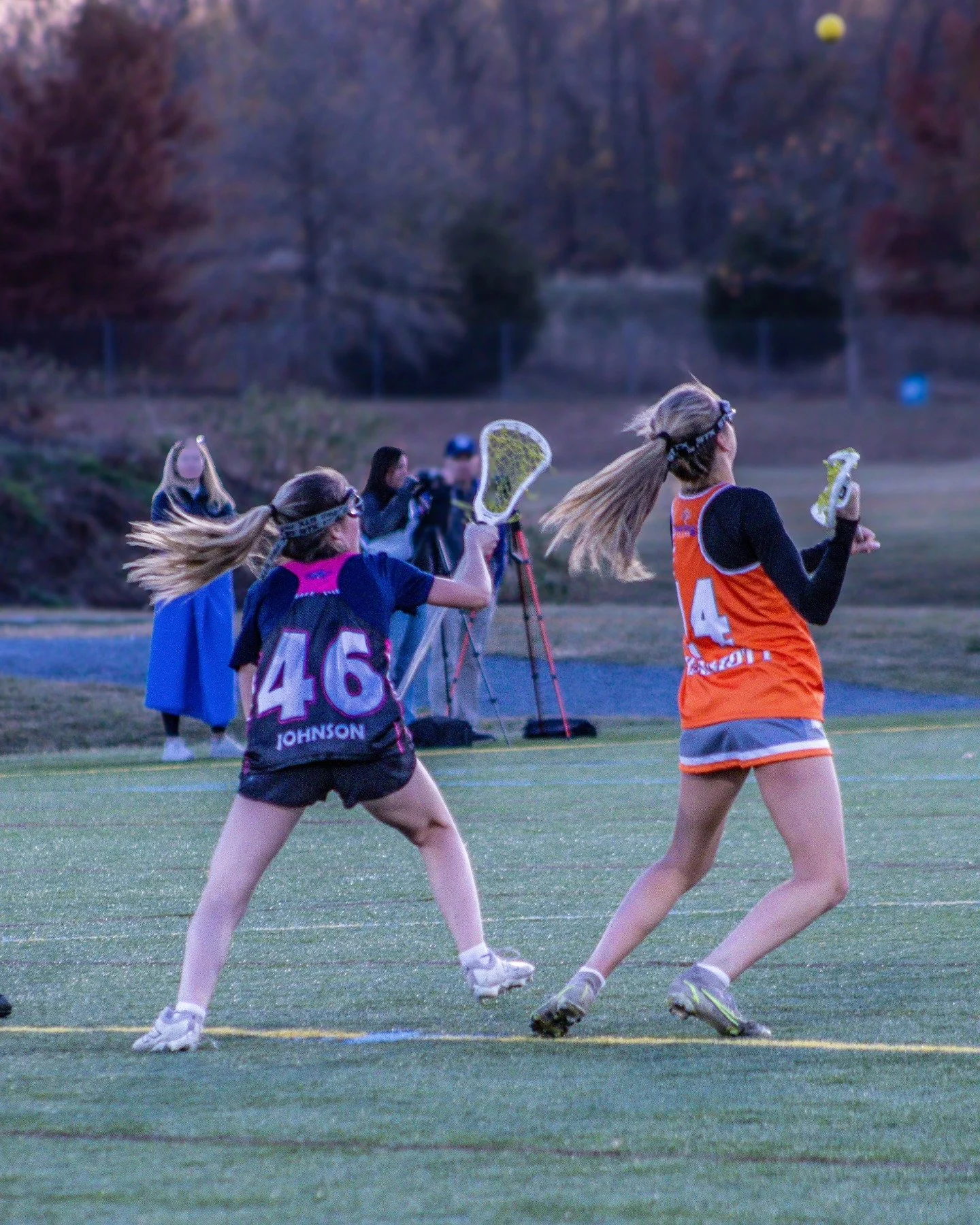 Sports photography Baltimore Maryland DMV. Two young women playing lacrosse on a field during dusk, with one in a black and pink jersey and the other in an orange and black jersey, both holding lacrosse sticks.