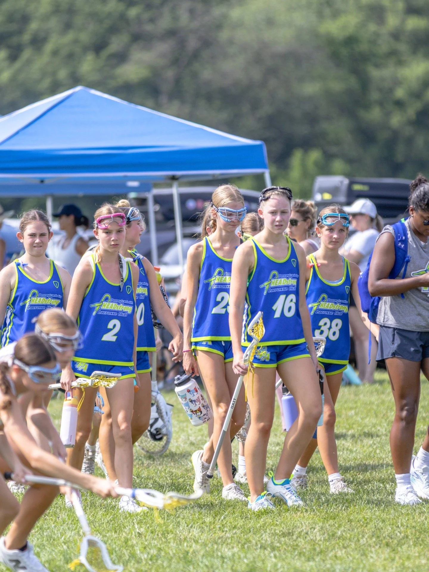 Sports photography Baltimore Maryland DMV. A group of young women in blue and yellow lacrosse uniforms walking on a grassy field with water bottles, tents, and vehicles in the background.
