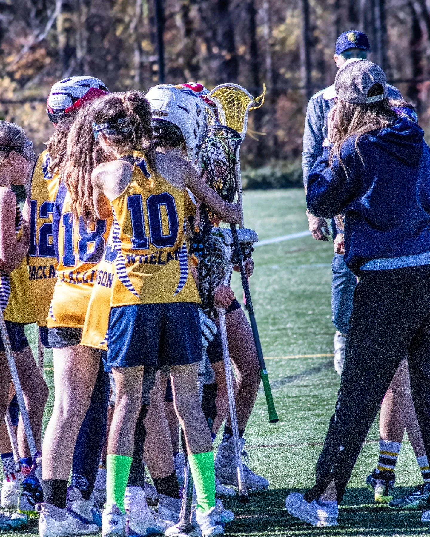 Sports photography Baltimore Maryland DMV. Girls' lacrosse team huddled on the field around their coach during a game. The players are in uniforms with yellow jerseys and blue numbers, holding lacrosse sticks. 