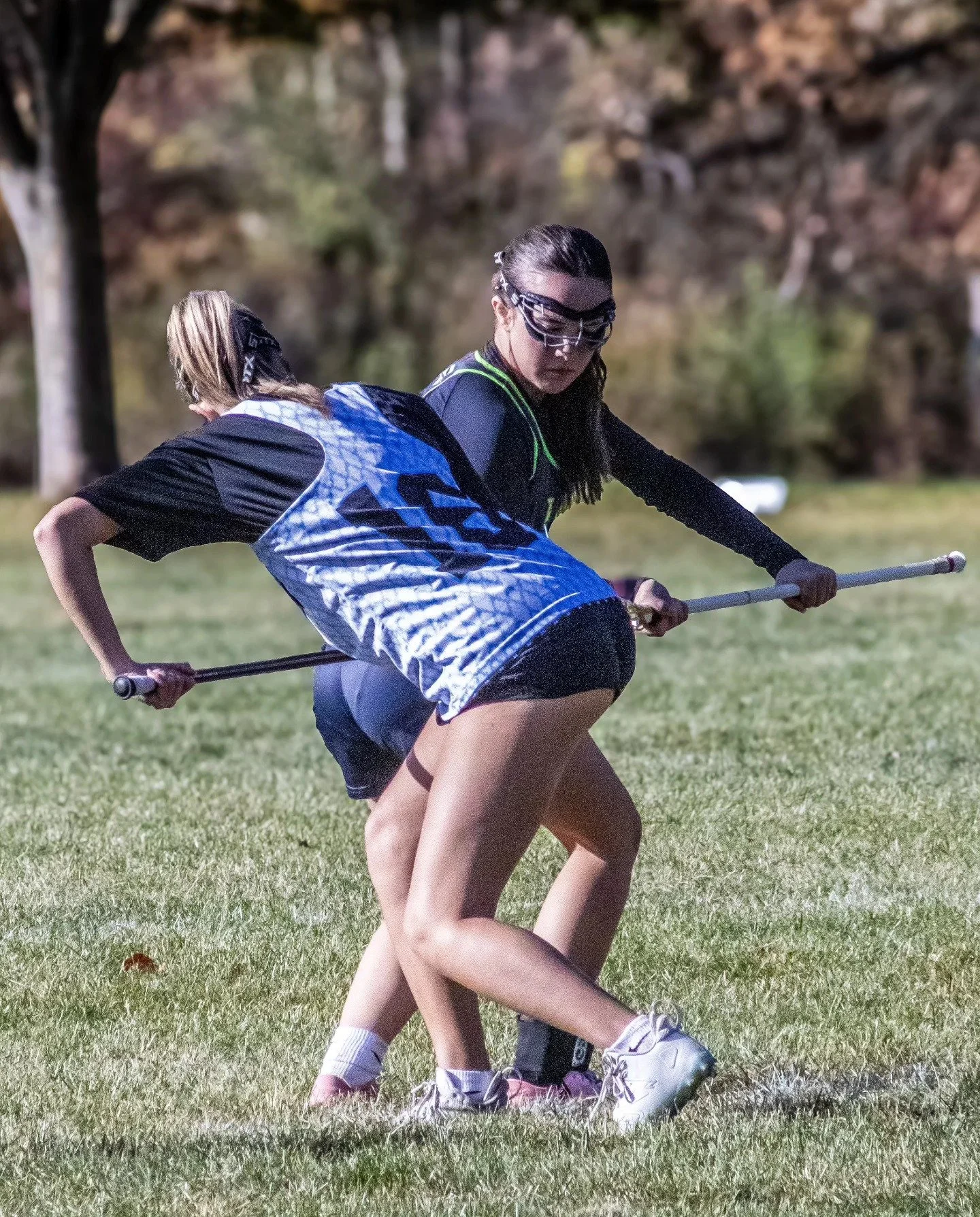 Sports photography Baltimore Maryland DMV. Two young women playing field hockey on a grassy field, wearing sports uniforms, goggles, and holding hockey sticks.