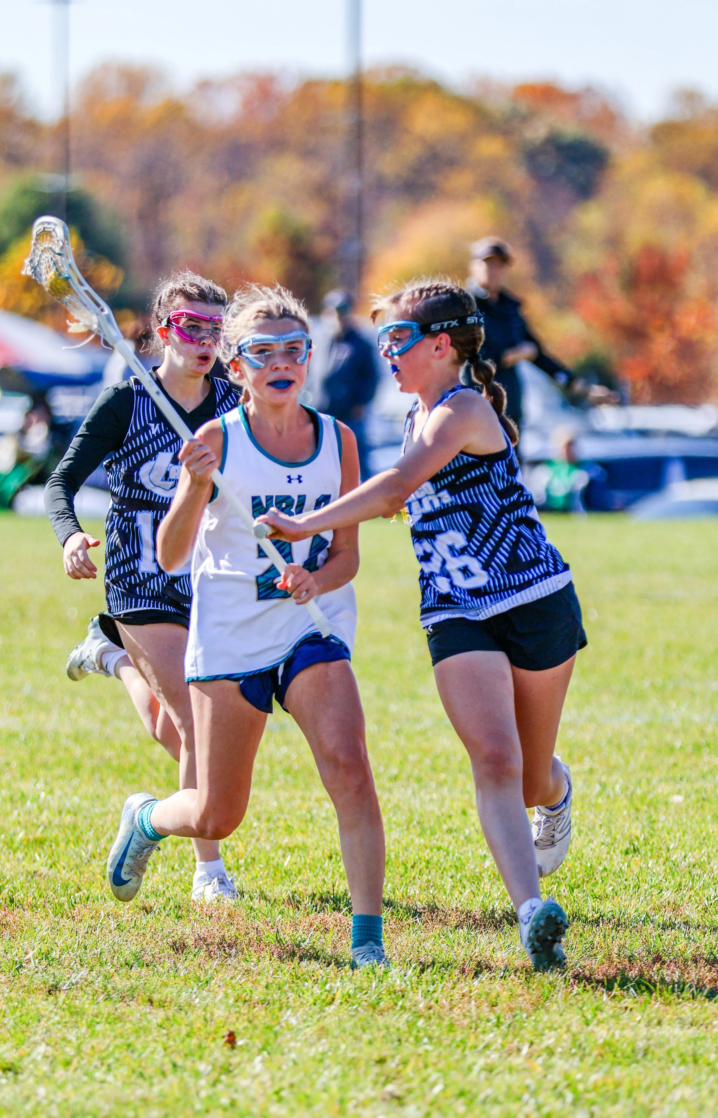 Sports photography Baltimore Maryland DMV. Young female lacrosse players running on a grassy field during daytime, wearing sports gear and goggles, with a background of autumn-colored trees.