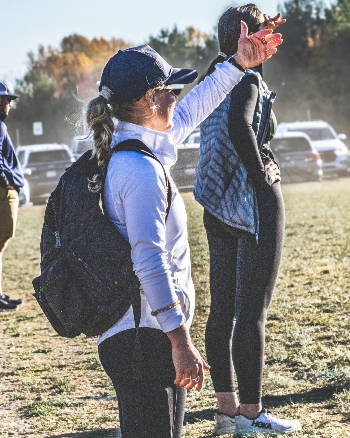 Sports photography Baltimore Maryland DMV. People standing outdoors in athletic clothing, some with backpacks, engaged in conversation or watching something, with cars parked in the background and trees with fall foliage.