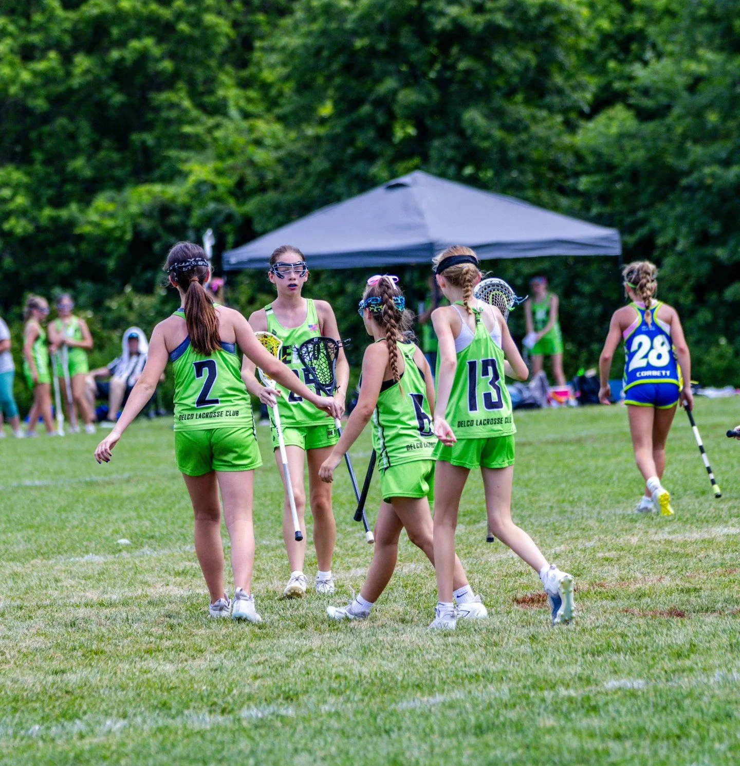 Sports photography Baltimore Maryland DMV. Group of young girls playing lacrosse on a grassy field, wearing green uniforms with numbers and carrying lacrosse sticks, with trees and a tent in the background.