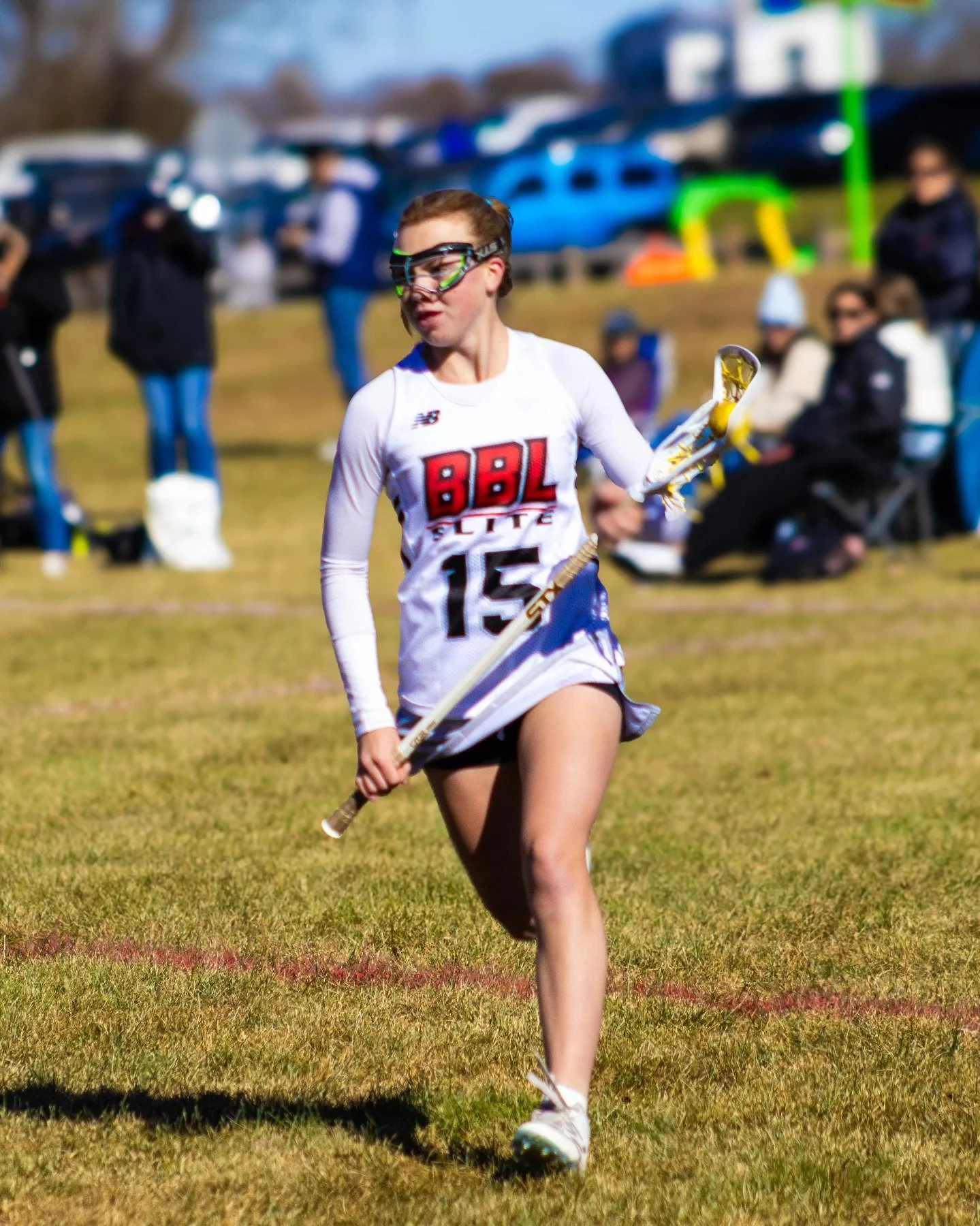 Sports photography Baltimore Maryland DMV. A young female athlete running on a grassy field with a lacrosse stick in her hand, wearing a white sports jersey with the number 15 and protective goggles, with spectators and parked cars in the background.