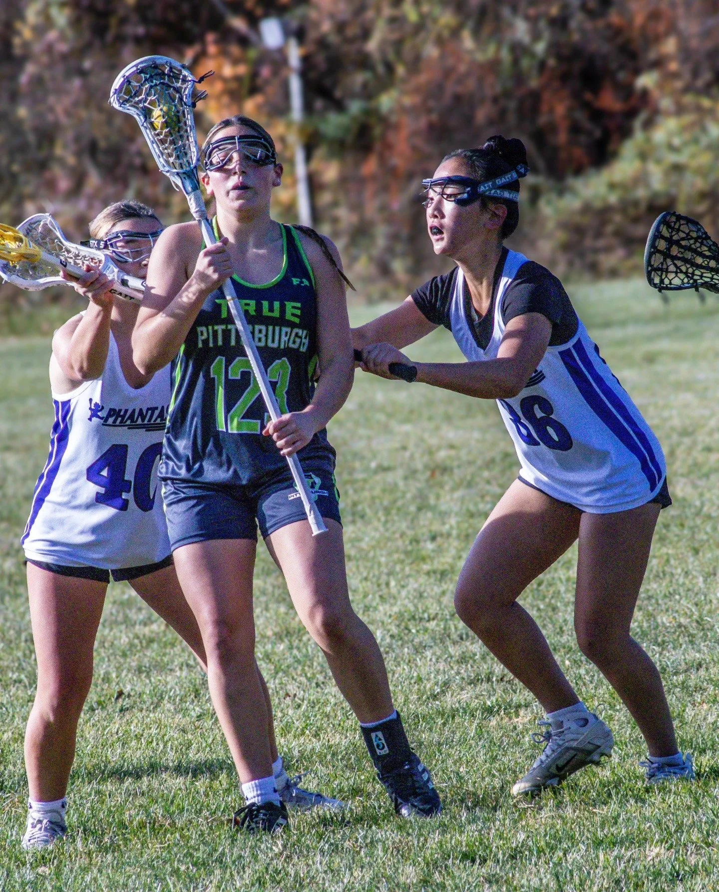 Sports photography Baltimore Maryland DMV. Four young women playing lacrosse on a grassy field, wearing sports jerseys, protective goggles, and holding lacrosse sticks.