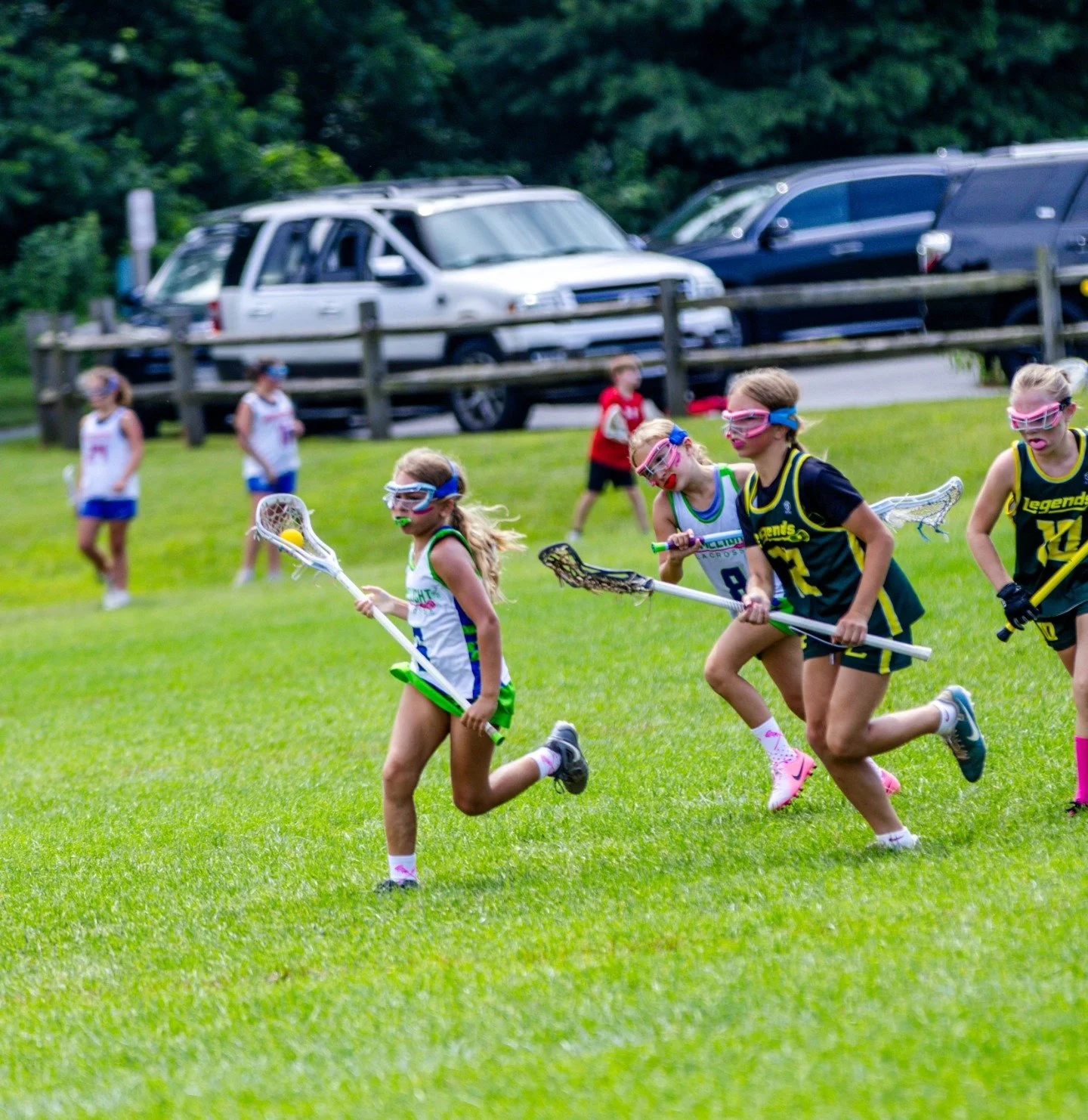 Sports photography Baltimore Maryland DMV. Young girls playing lacrosse on a grassy field, wearing protective gear and colored uniforms.